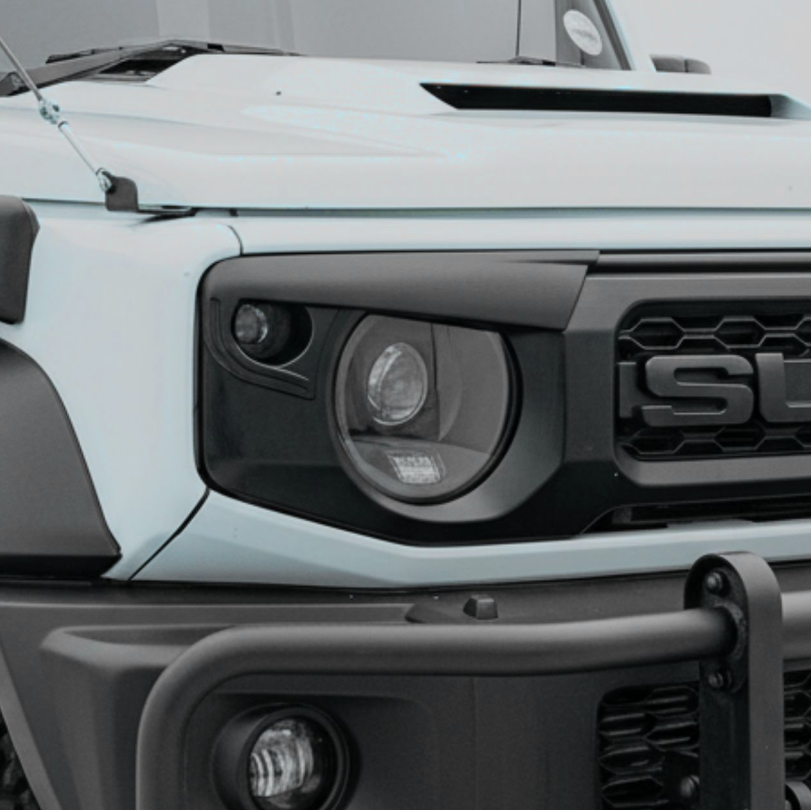 Close-up of the front of a white and black Land Rover Defender, showing the headlight, grille, and a windshield with a sticker.
