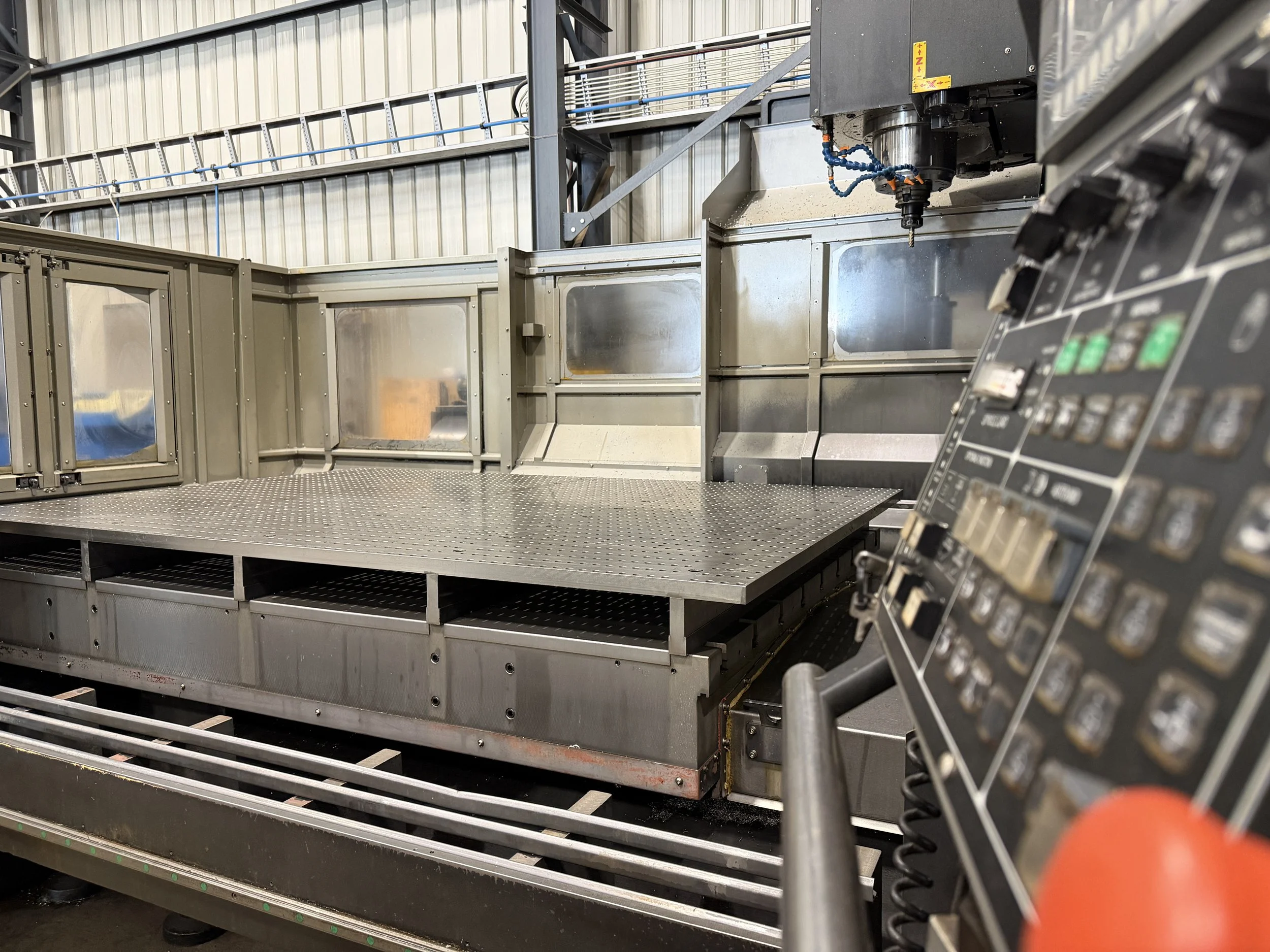 An industrial machine with a metal platform, control panel with buttons and switches, and a robotic arm with a blue flexible tube. The machine is inside a warehouse with metal walls and a staircase nearby.