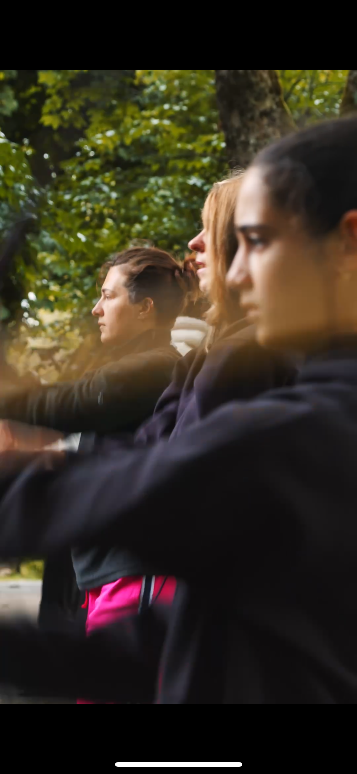 Trois femmes assises côte à côte sous un arbre, ayant l'air concentré ou pensif, vêtues de vêtements décontractés, dans un environnement naturel avec des feuilles vertes en arrière-plan.