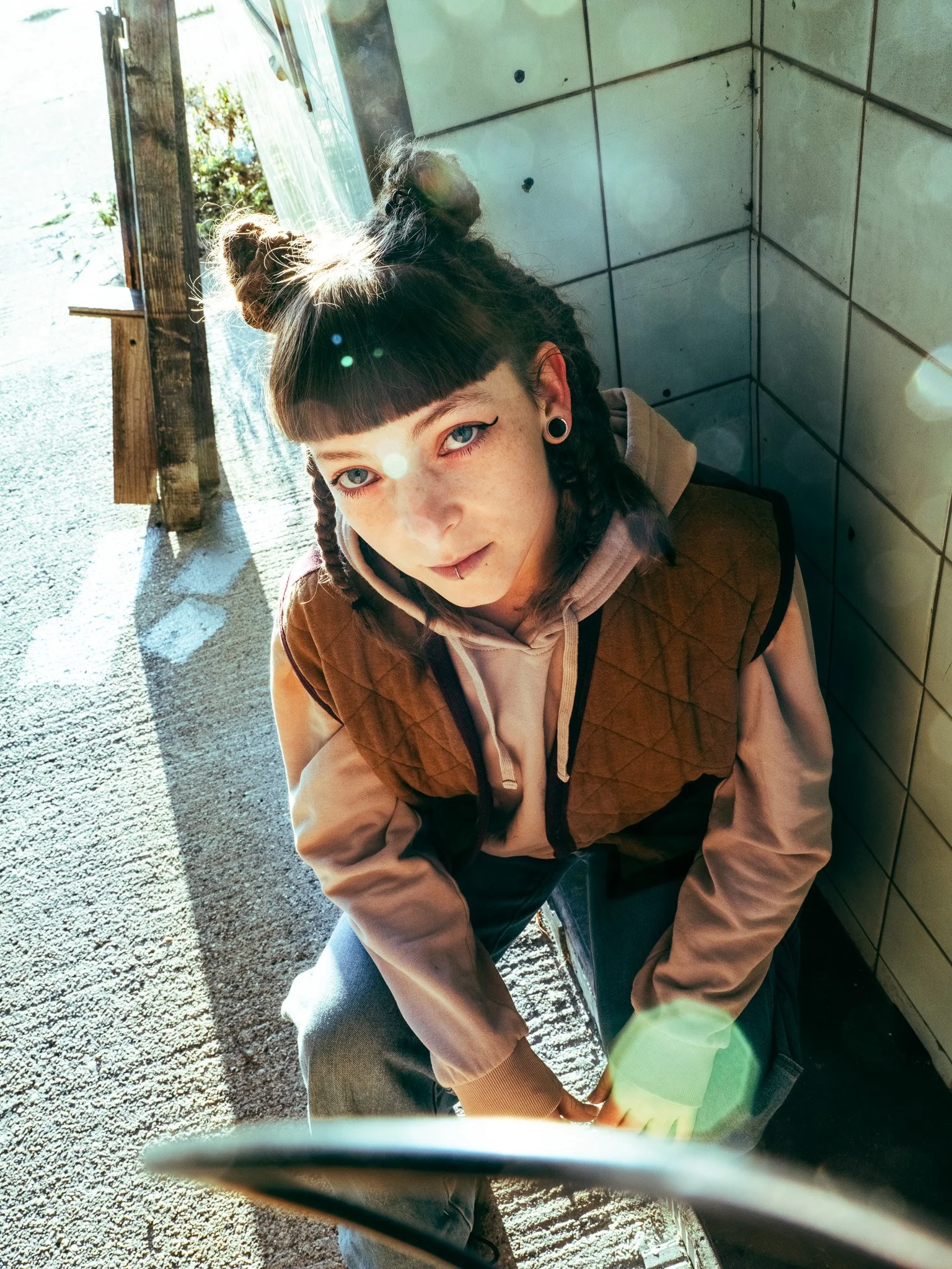 Une jeune femme aux cheveux bruns coiffés en deux buns, portant un hoodie beige et une veste brune avec des épaules dénudées, assise près d'une porte ouverte, regardant vers la caméra, avec un fond en extérieur et un mur en carrelage gris derrière elle.