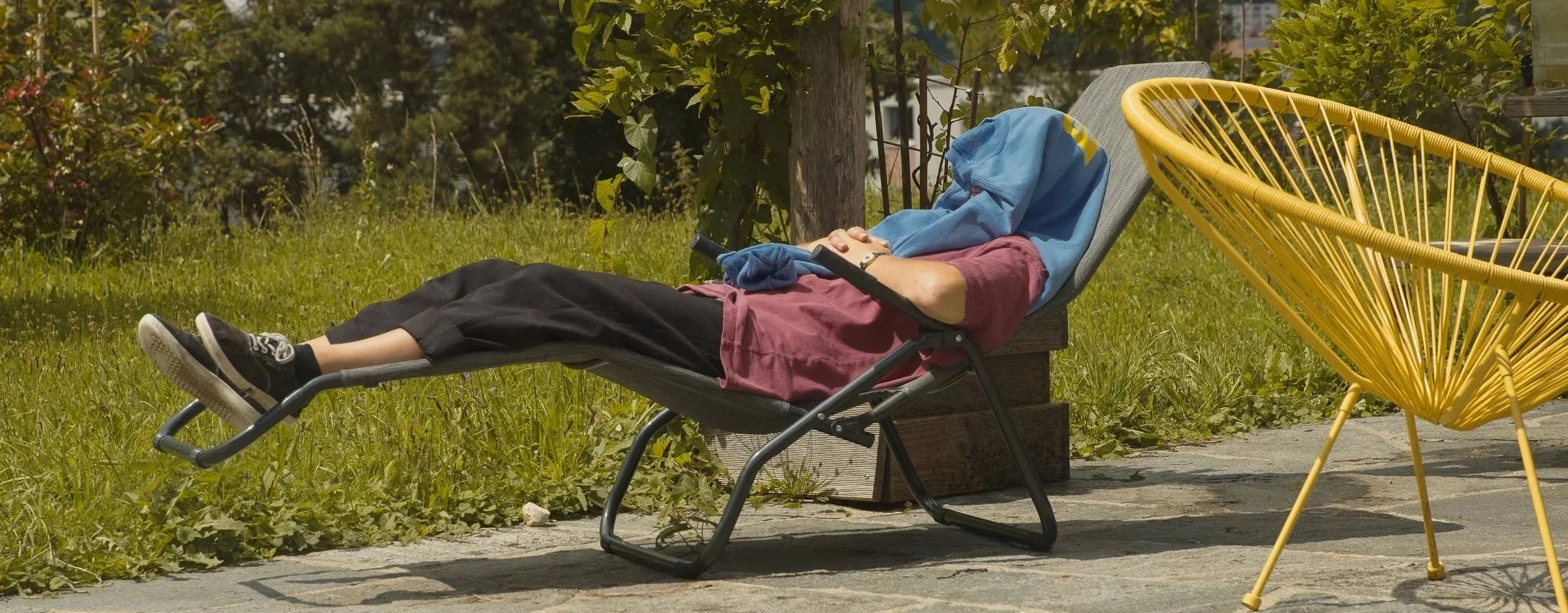 Personne allongée sur un fauteuil de jardin, tête et visage couverts par un t-shirt, tenant une manette de jeu, dans un espace extérieur avec herbe et arbres.