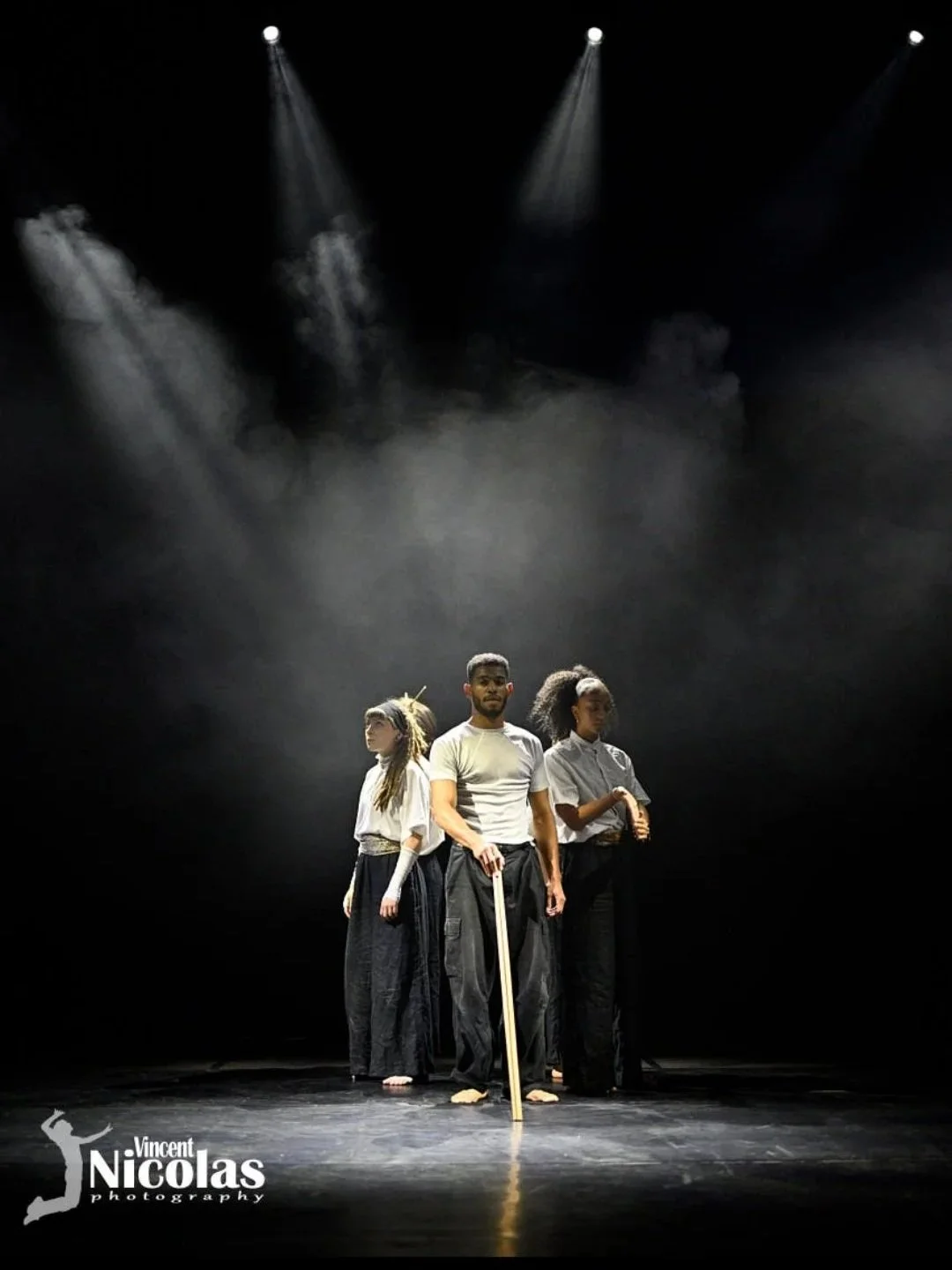 Trois personnes, deux femmes et un homme, debout sur une scène sombre sous des projecteurs, portant des vêtements décontractés et posant sérieusement.