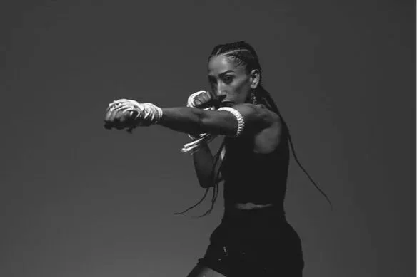 Femme boxeuse en posture de combat, portant des gants de boxe, vêtue d'une tenue athlétique noire, dans un décor sombre.