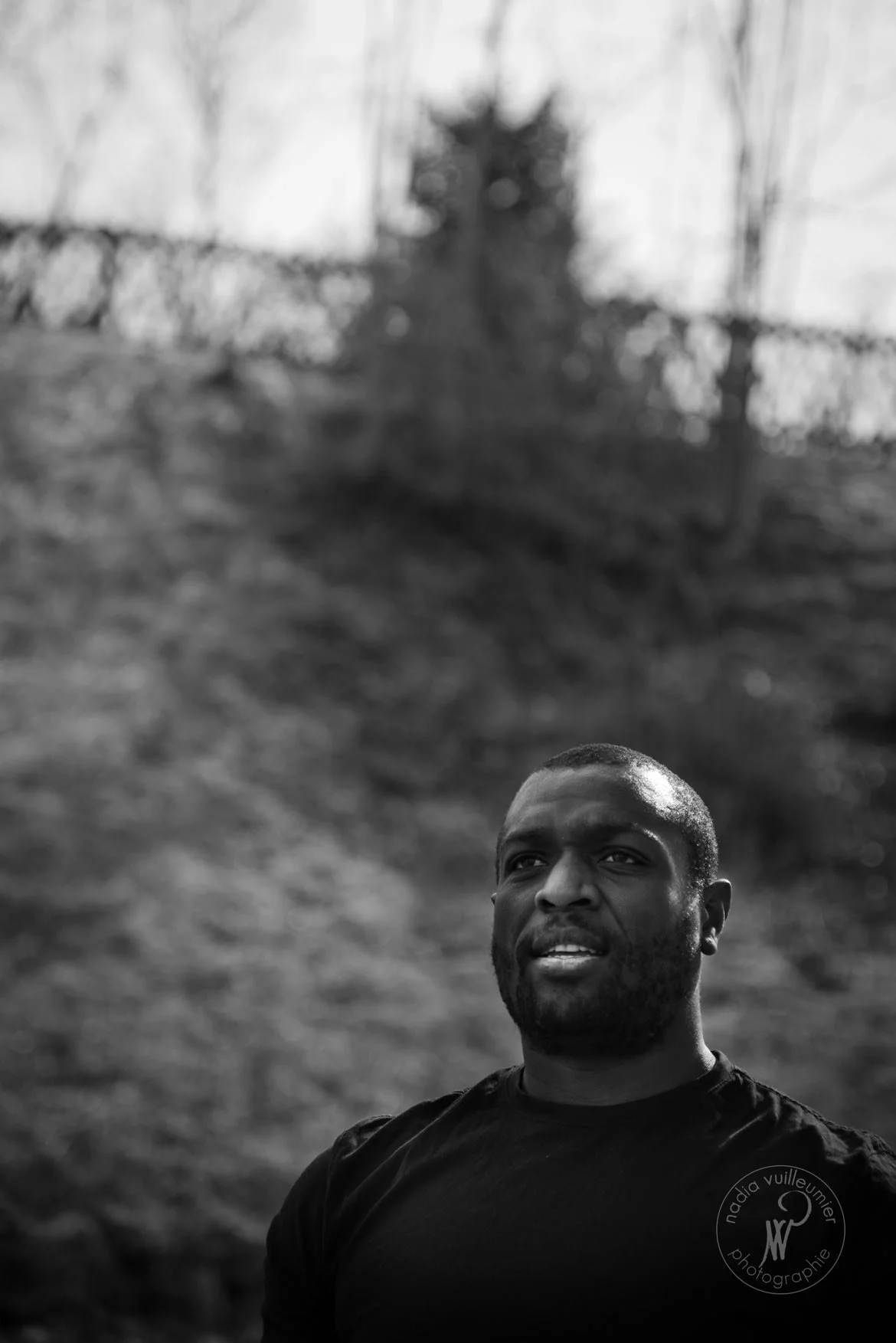 Portrait d'un homme noir à la peau mate, avec une expression neutre, portant un t-shirt noir, en extérieur, avec des arbres et une clôture flous en arrière-plan, en noir et blanc.