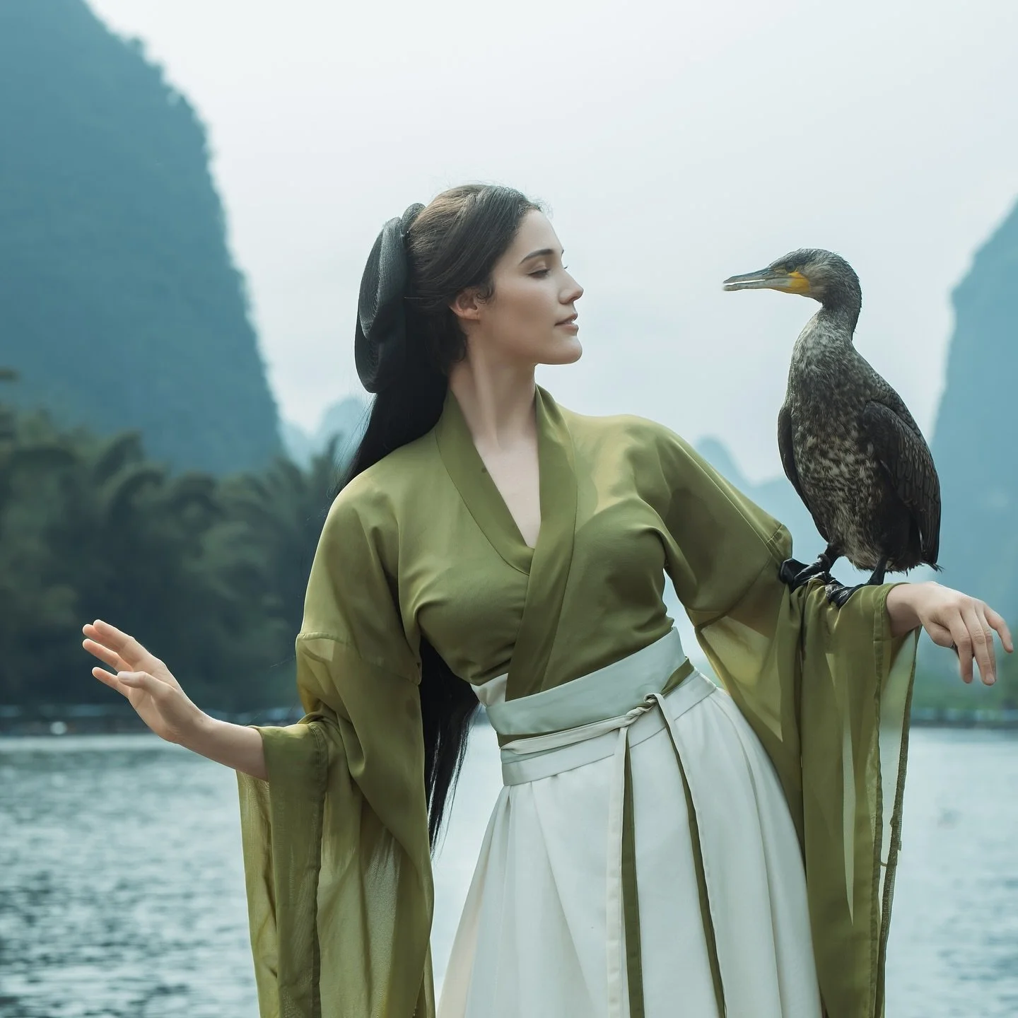 Une femme vêtue d'une robe vert olive et d'une jupe blanche, avec un faisan posé sur son bras, se tient près d'une rivière entourée de montagnes et de végétation.