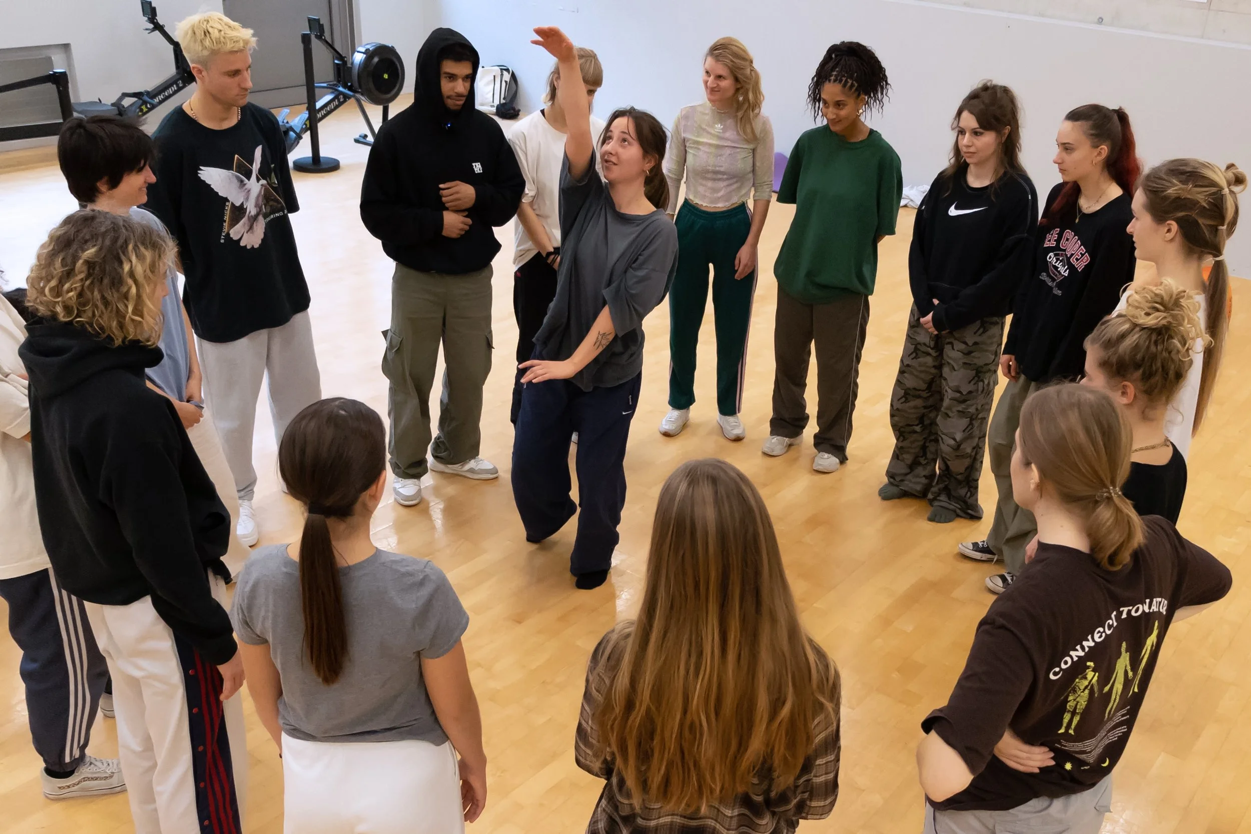 Groupe de jeunes personnes en cercle dans une salle de studio ou de gym, certaines en train de parler ou d'écouter, participation active à une activité de groupe.