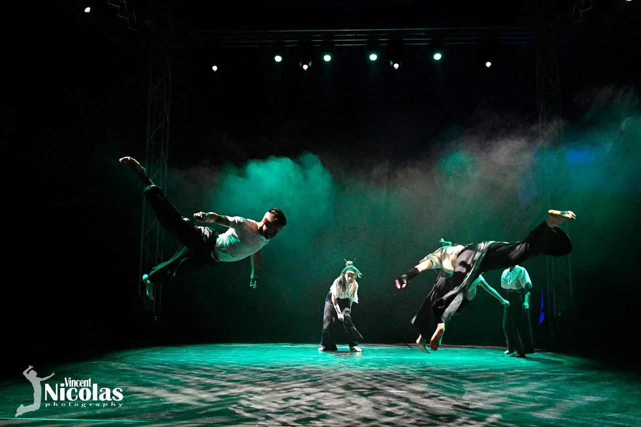 Des danseurs en costume effectuent une performance de danse contemporaine sur scène, éclairés par des lumières vertes et bleues.