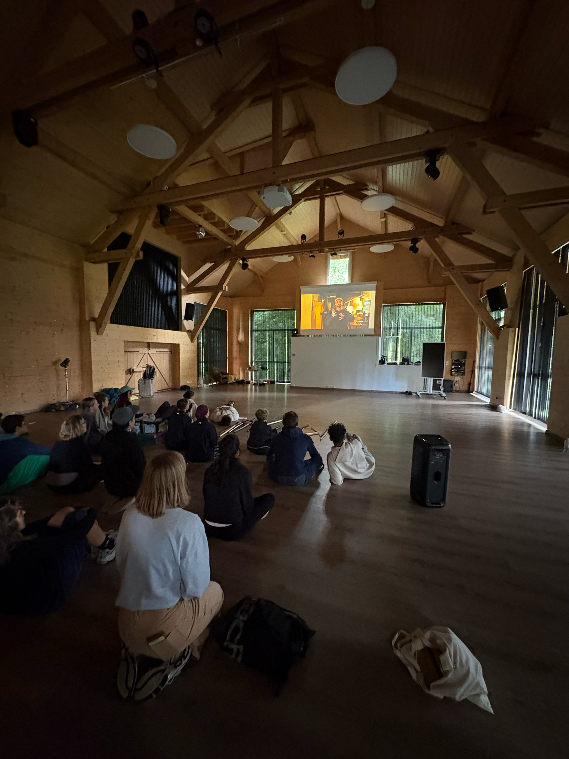 Groupe de personnes assises sur le sol dans une grande salle en bois, regardant un écran de projection au fond, dans un environnement chaleureux avec de grandes fenêtres et un plafond en poutres.
