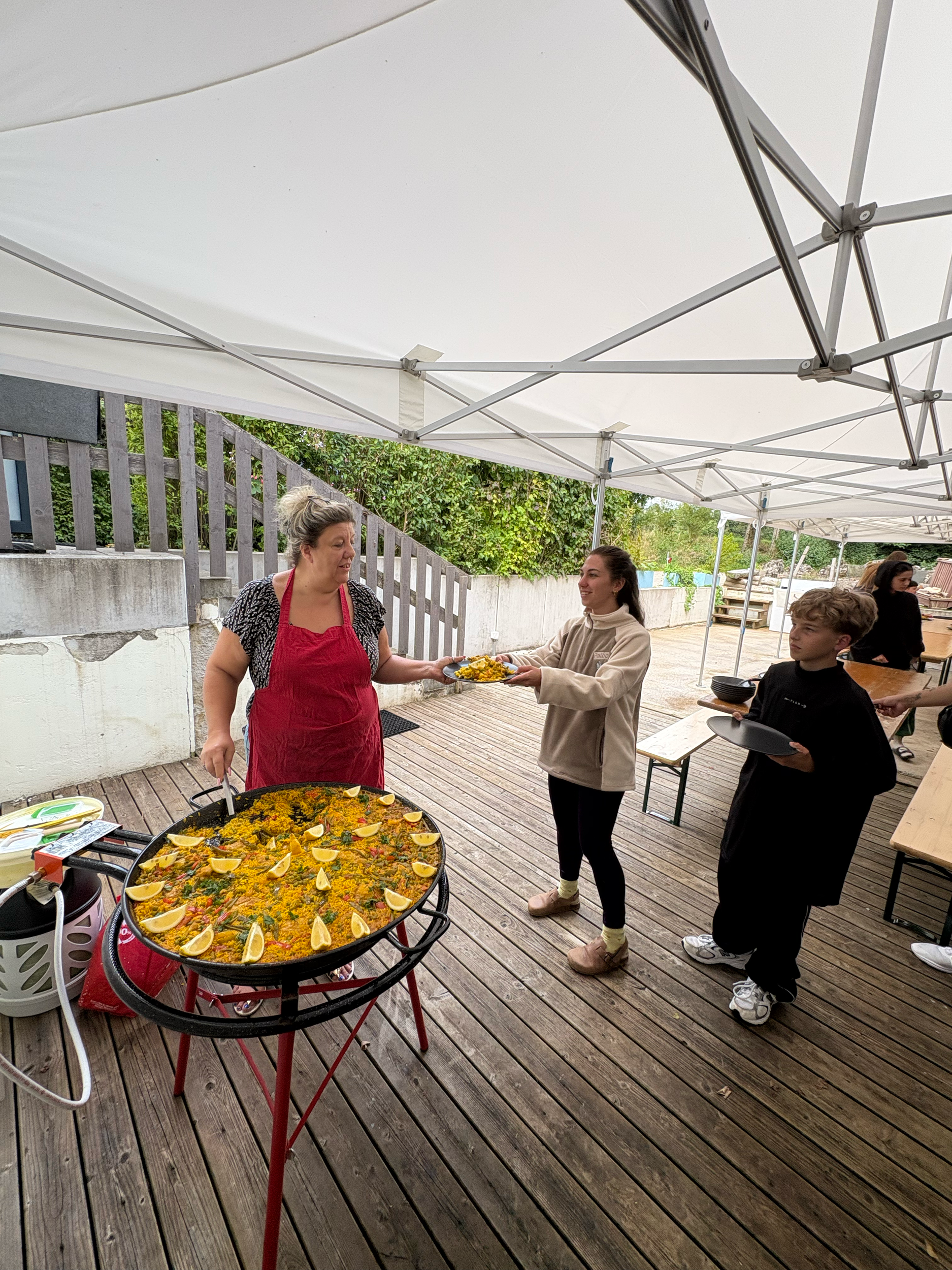 Une femme en chemise à manches longues claire et un jean reçoit un plat de nourriture d'une autre femme portant un tablier rouge. Il y a une grande paella garnie de citrons coupés en quartiers sur un support, sous une tente blanche en extérieur, avec