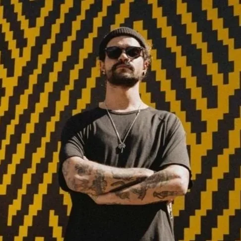 Homme avec lunettes de soleil, casquette, pendentif, tatouages sur les bras, debout devant un mur jaune et noir avec un motif géométrique.