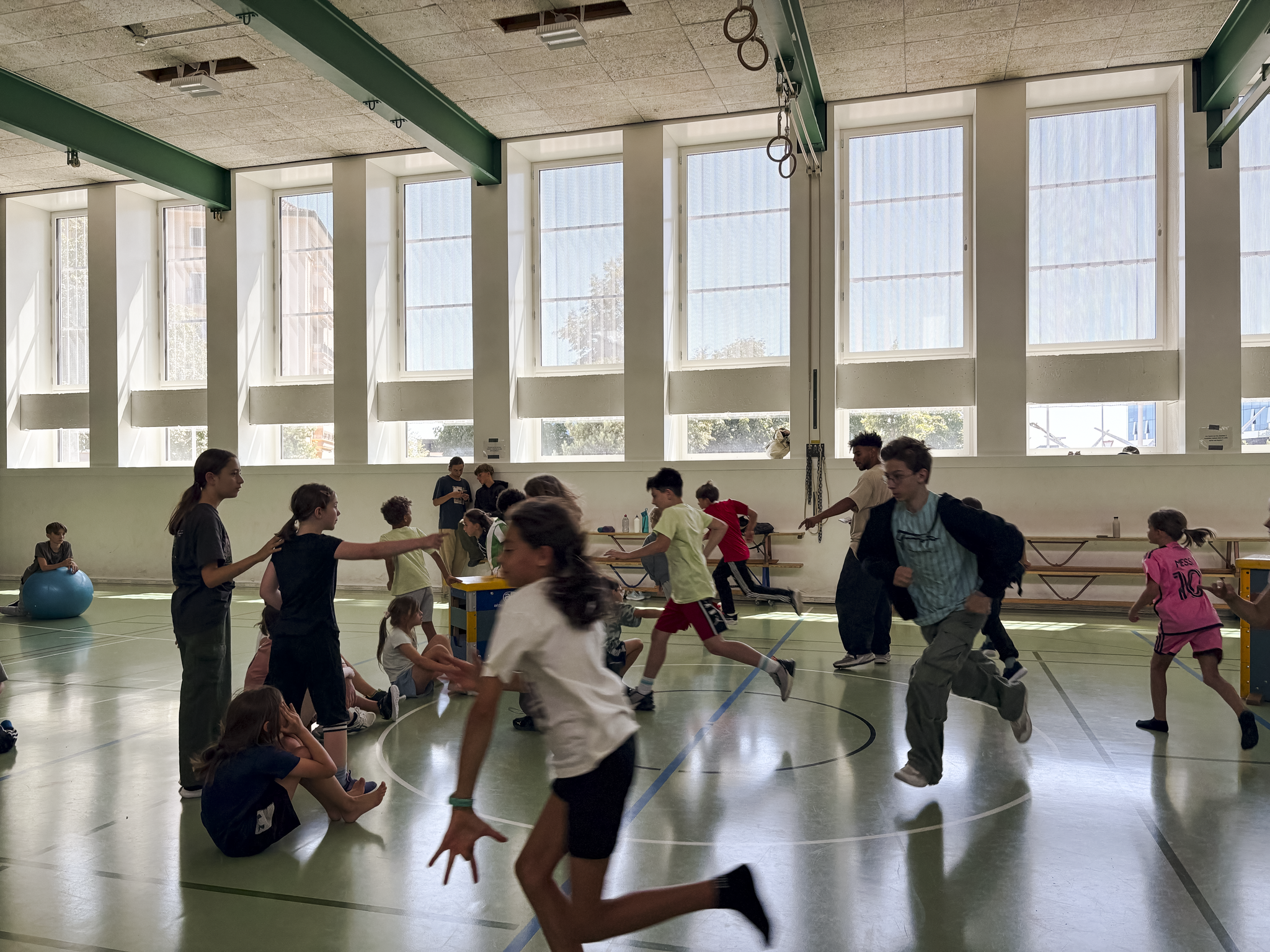 Une groupe d'enfants et d'adultes dans une salle de sport, certains jouant, d'autres assis ou se tenant debout, avec de grandes fenêtres laissant entrer la lumière naturelle.