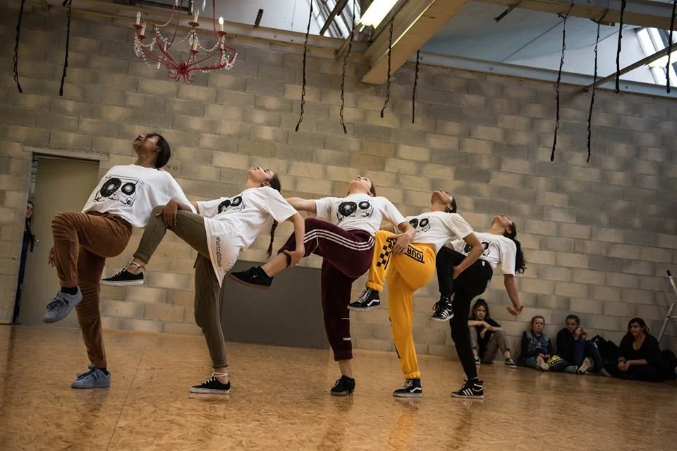 Cinq femmes dans une salle de danse exécutent une chorégraphie en équilibre sur une jambe, avec les bras et la tête inclinés en arrière. Elles portent toutes un t-shirt blanc avec un design de DJ et différentes couleurs de pantalons. Des personnes assises contre le mur regardent l'exercice.
