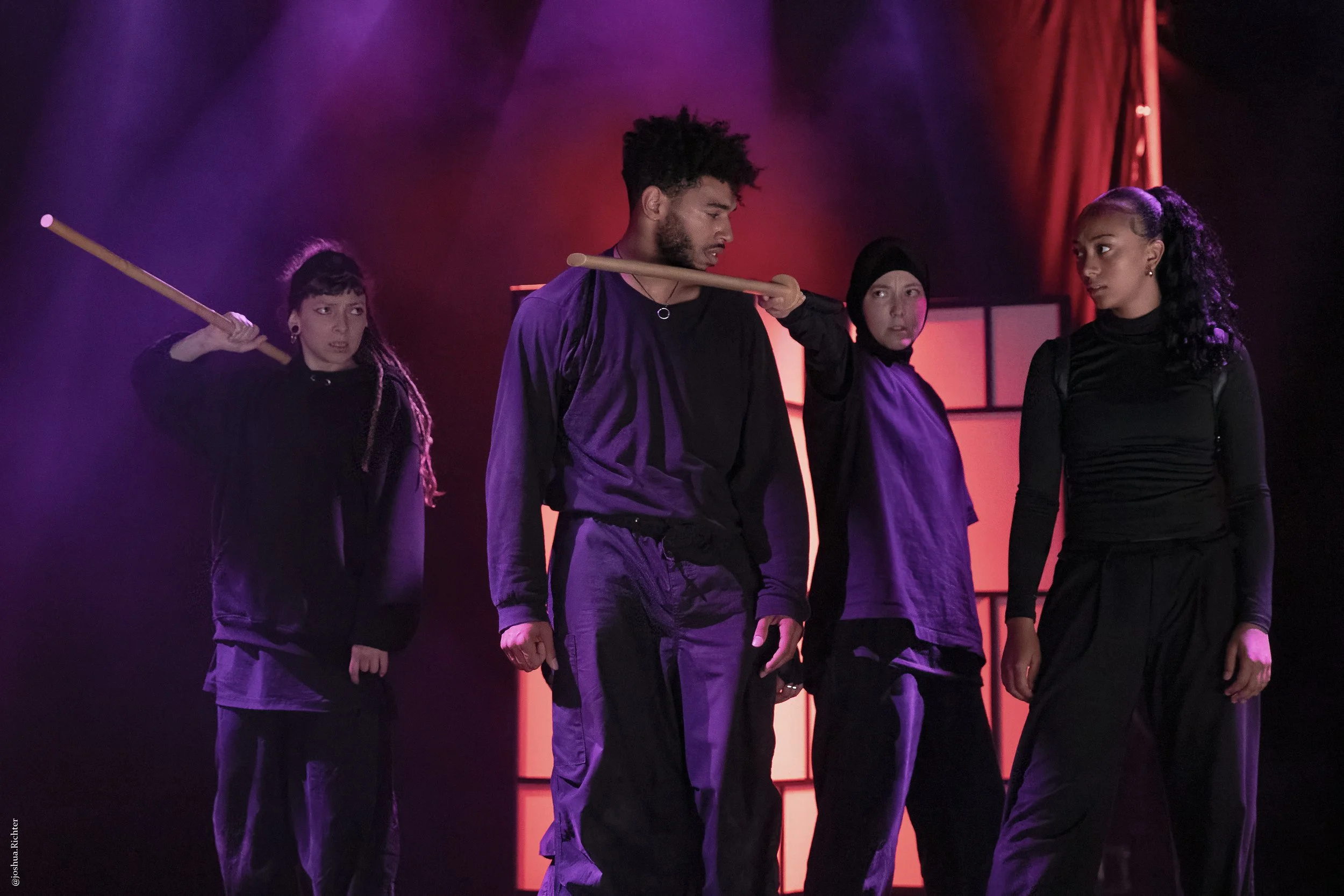 Quatre personnes en vêtements noirs sur scène avec des bâtons, dans une mise en scène dramatique avec éclairage rouge et violet.