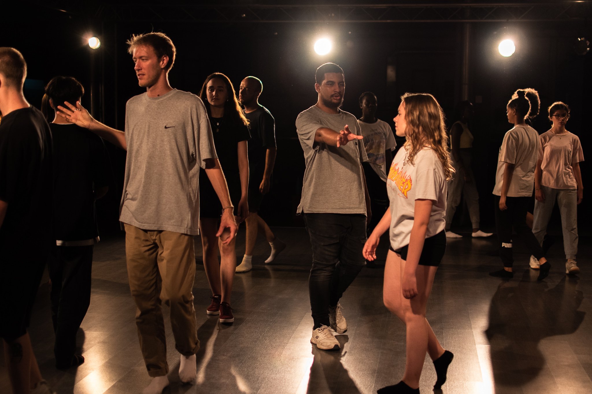 Groupe de jeunes dans une salle de danse avec éclairage au plafond, certains en mouvement, d'autres en position statique, échangeant des regards ou des gestes.