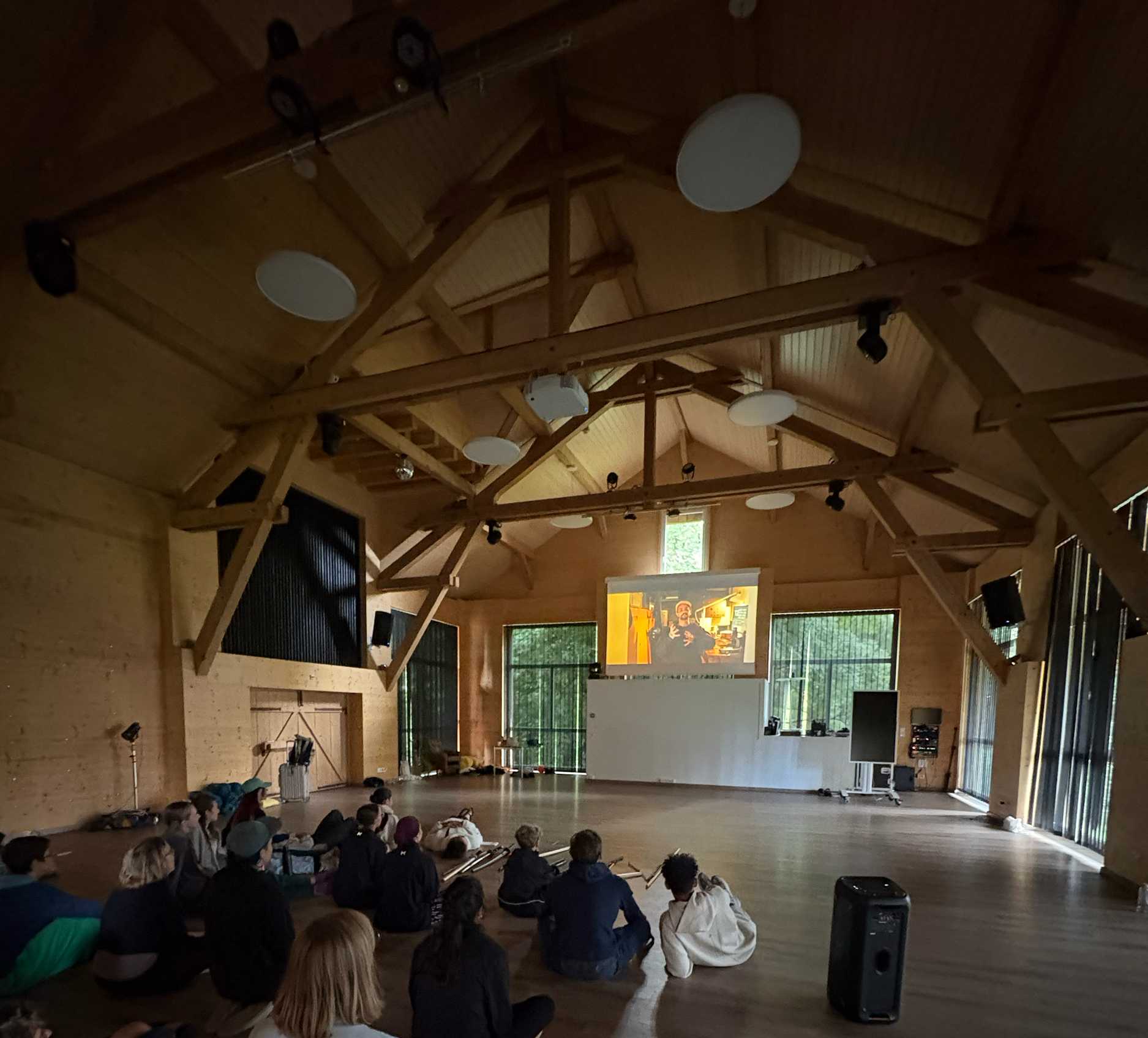 Groupe de personnes assises sur le sol d'une grande salle en bois regardant un écran projetant un film ou une présentation.