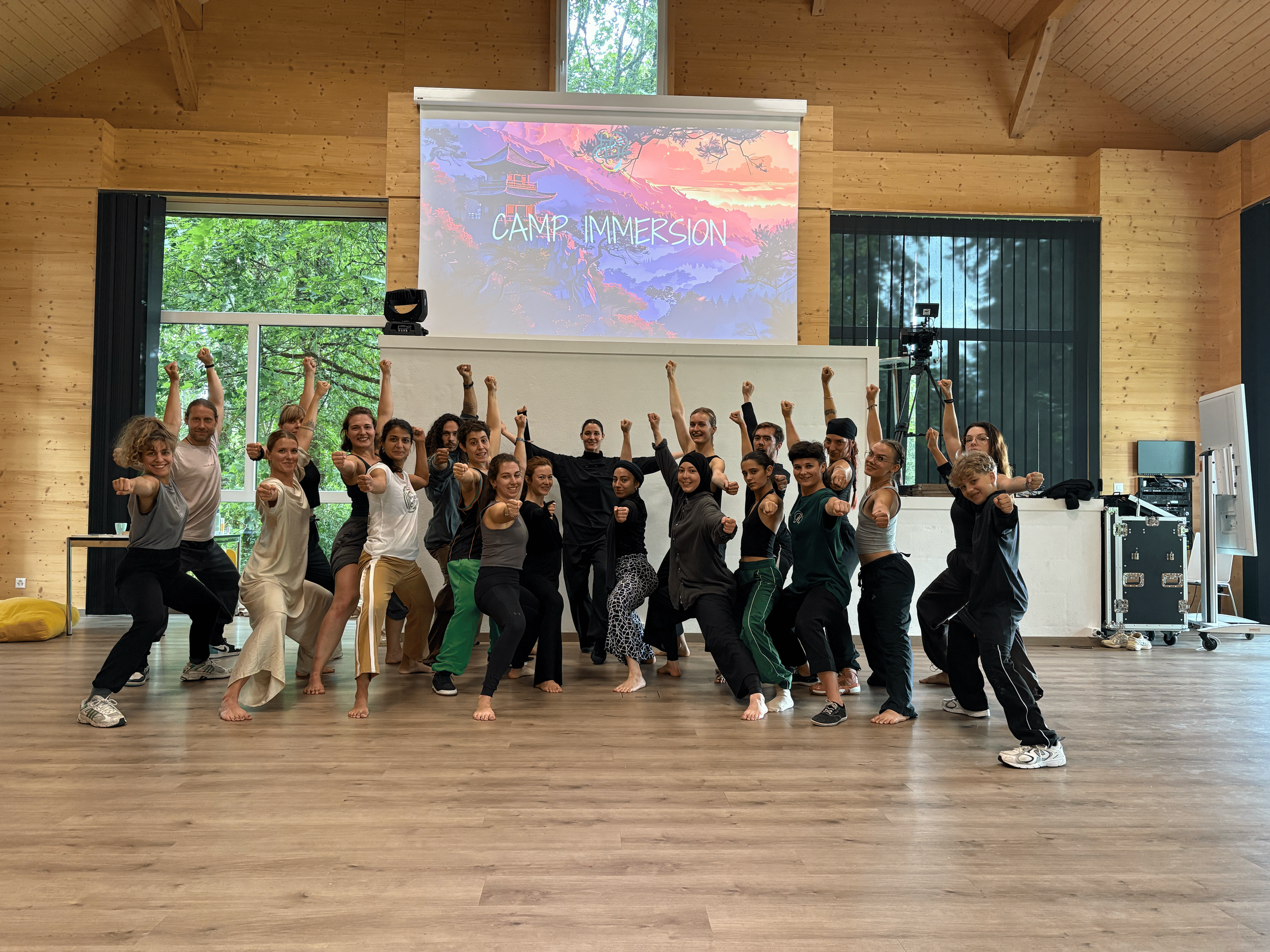 Groupe de personnes en position de combat sur un plancher en bois dans une salle avec murs en bois, derrière un écran avec l'inscription 'Camp Immersion' et une scène de nature.