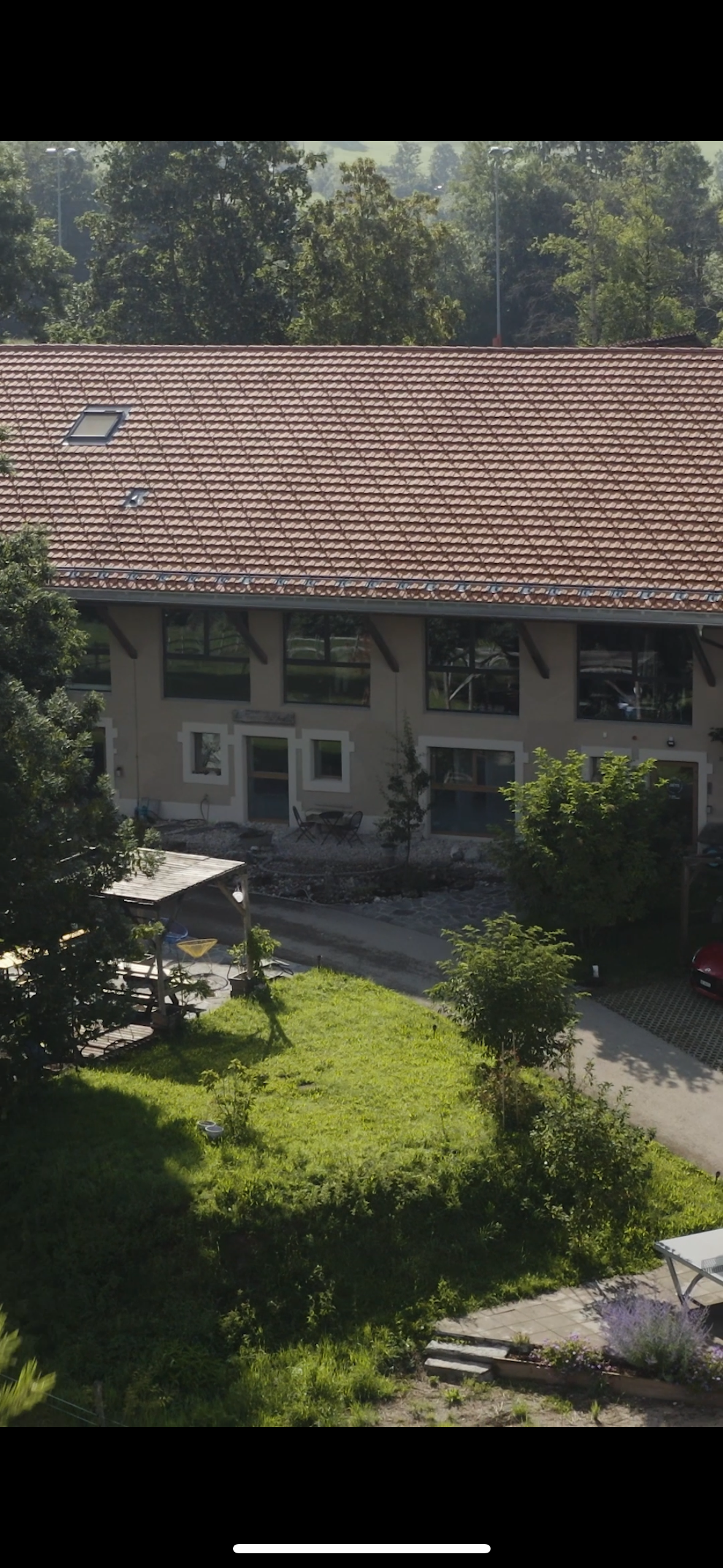 Une maison avec un toit en tuiles rouges, de nombreuses fenêtres, une petite terrasse, un jardin verdoyant avec des arbres et un espace de détente. La scène est en plein jour avec une lumière naturelle.
