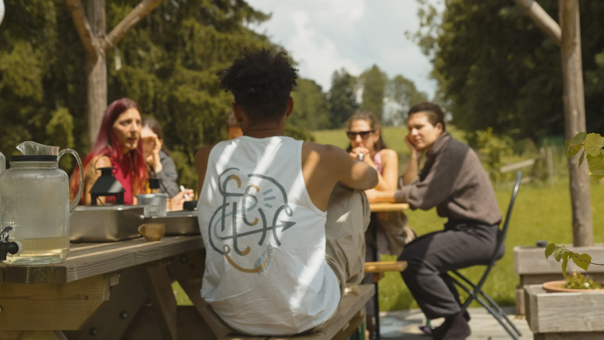 Groupe de jeunes personnes assises autour d'une table en plein air lors d'une journée ensoleillée dans un environnement naturel.
