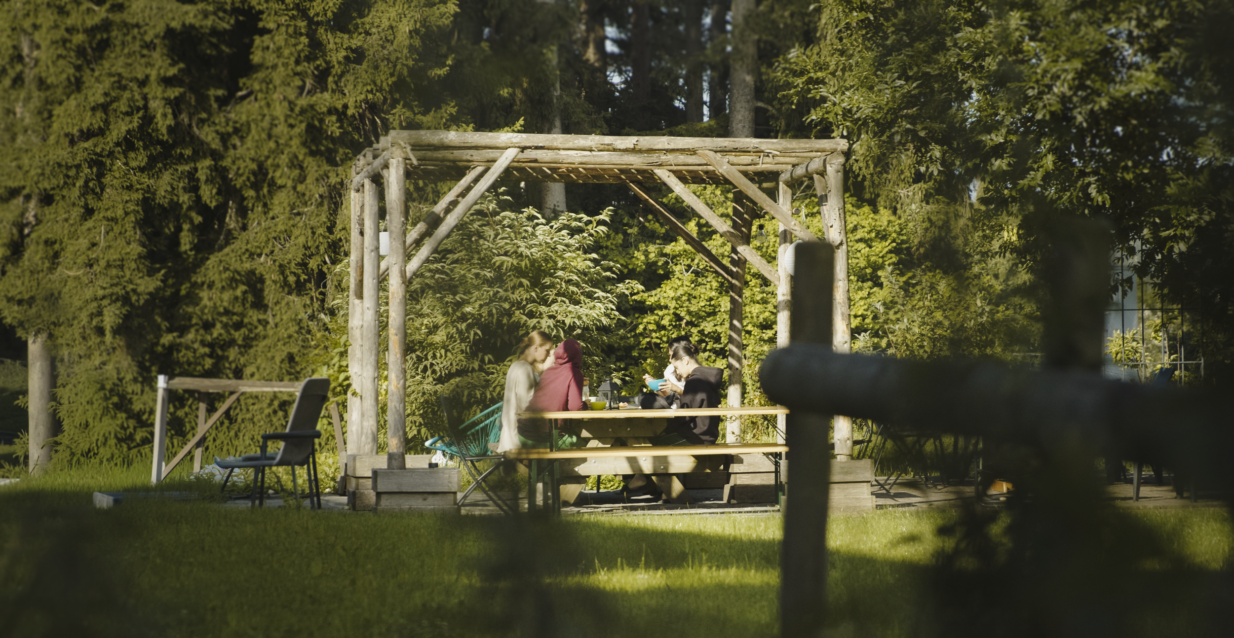 Trois personnes assises à une table en plein air sous une structure en bois, entourées d'arbres verts, en train de discuter ou de regarder leur téléphone.