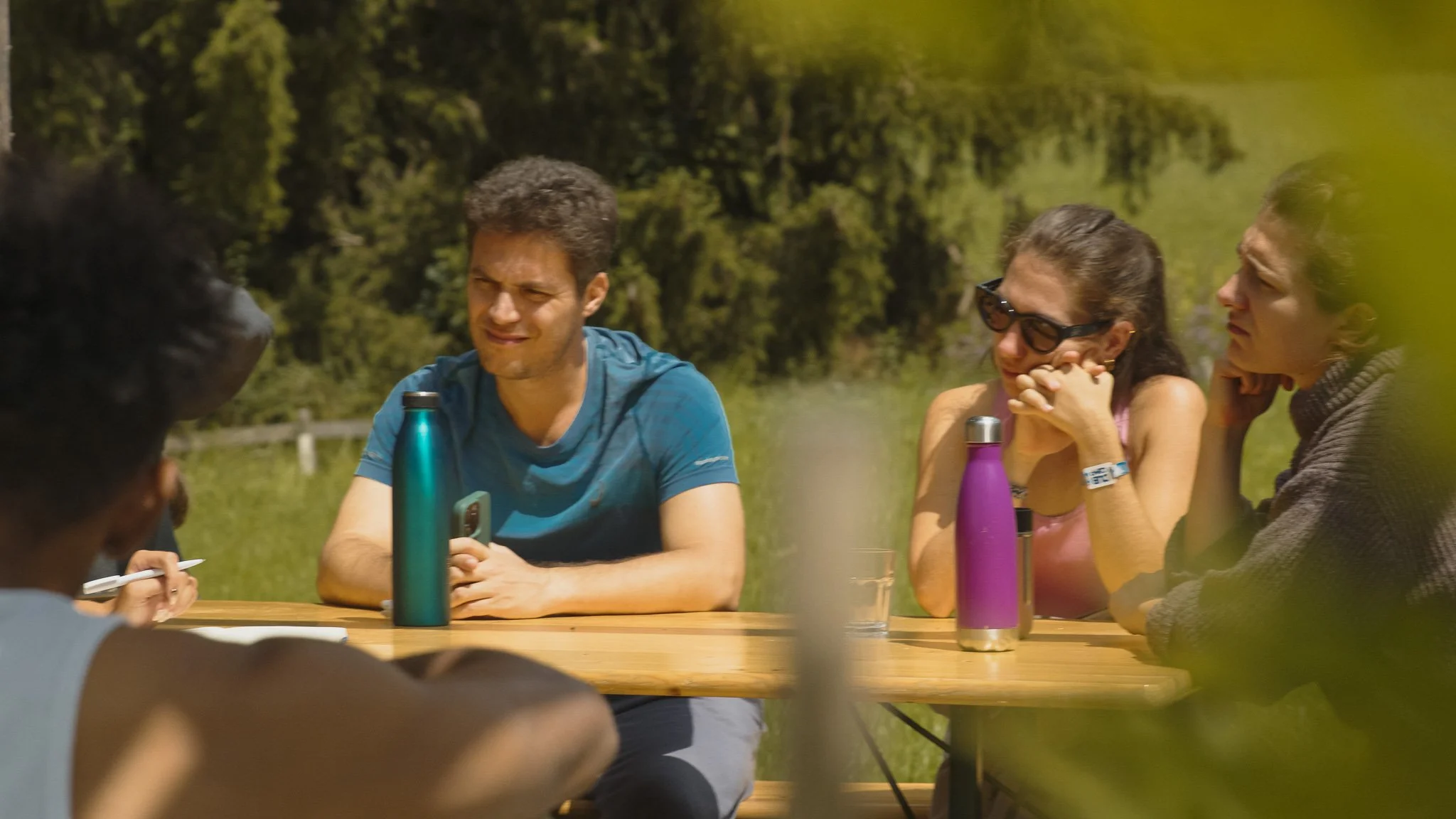 Groupe de personnes assises autour d'une table en plein air, discutant lors d'une journée ensoleillée dans un parc ou un espace naturel verdoyant.