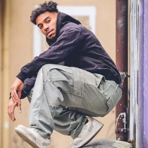 Jeune homme assis en équilibre sur un trottoir, portant un sweat à capuche noir, des pantalons cargo gris et des baskets blanches, dans un cadre urbain.