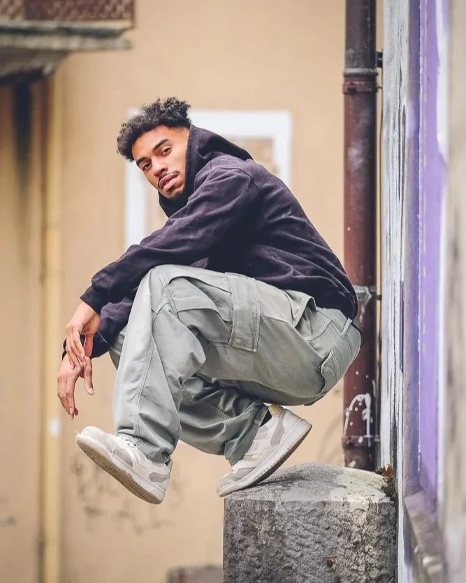 Jeune homme portant un sweat noir, des pantalons cargo gris, et des baskets blanches, assis en position accroupie sur un rebord en pierre, dans une rue urbaine.