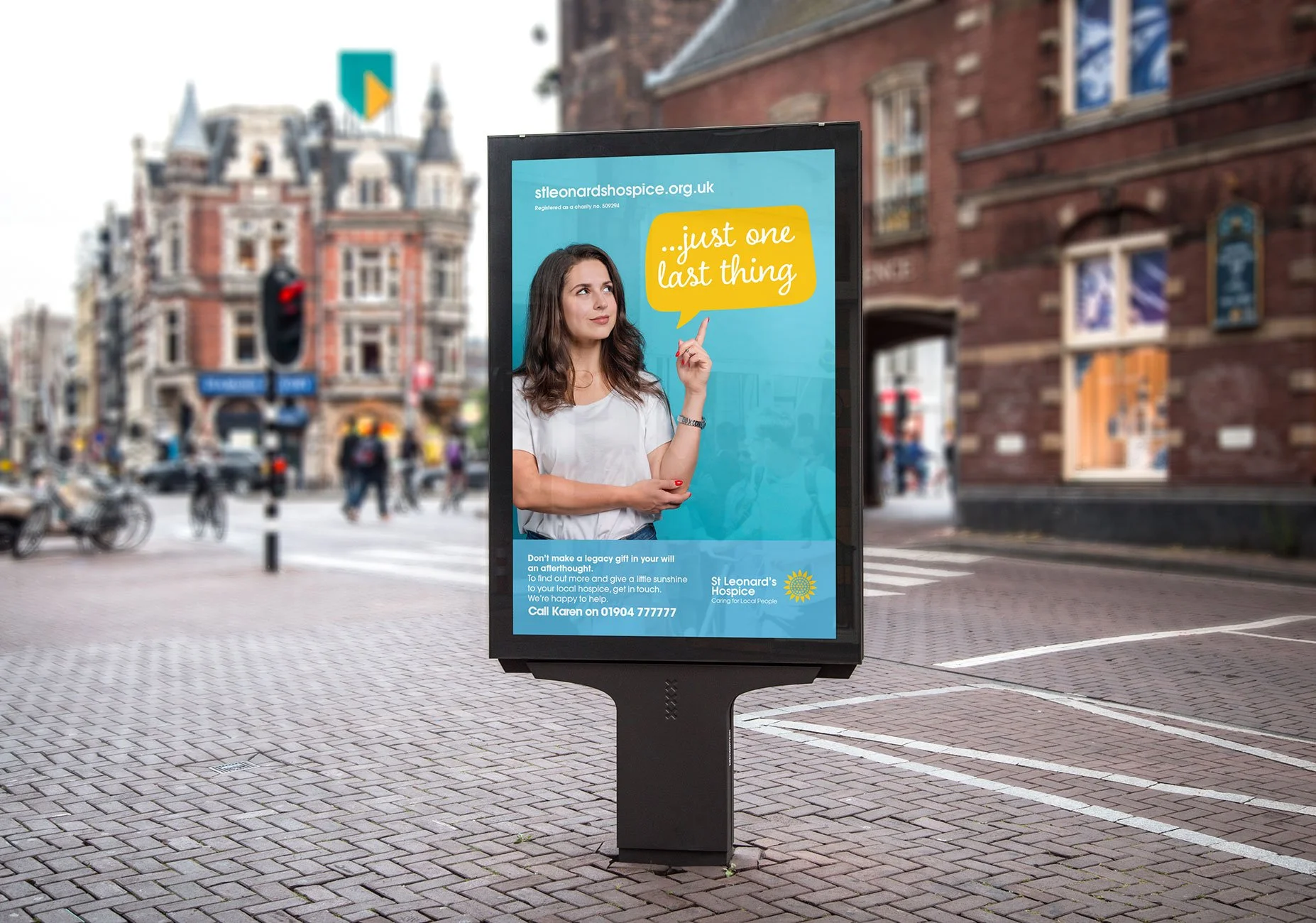 An outdoor advertisement for St. Leonard's Hospice featuring a woman with brown hair pointing upward with her index finger. The background of the ad is blue with a yellow speech bubble saying 'just one last thing.' There is additional text at the bot