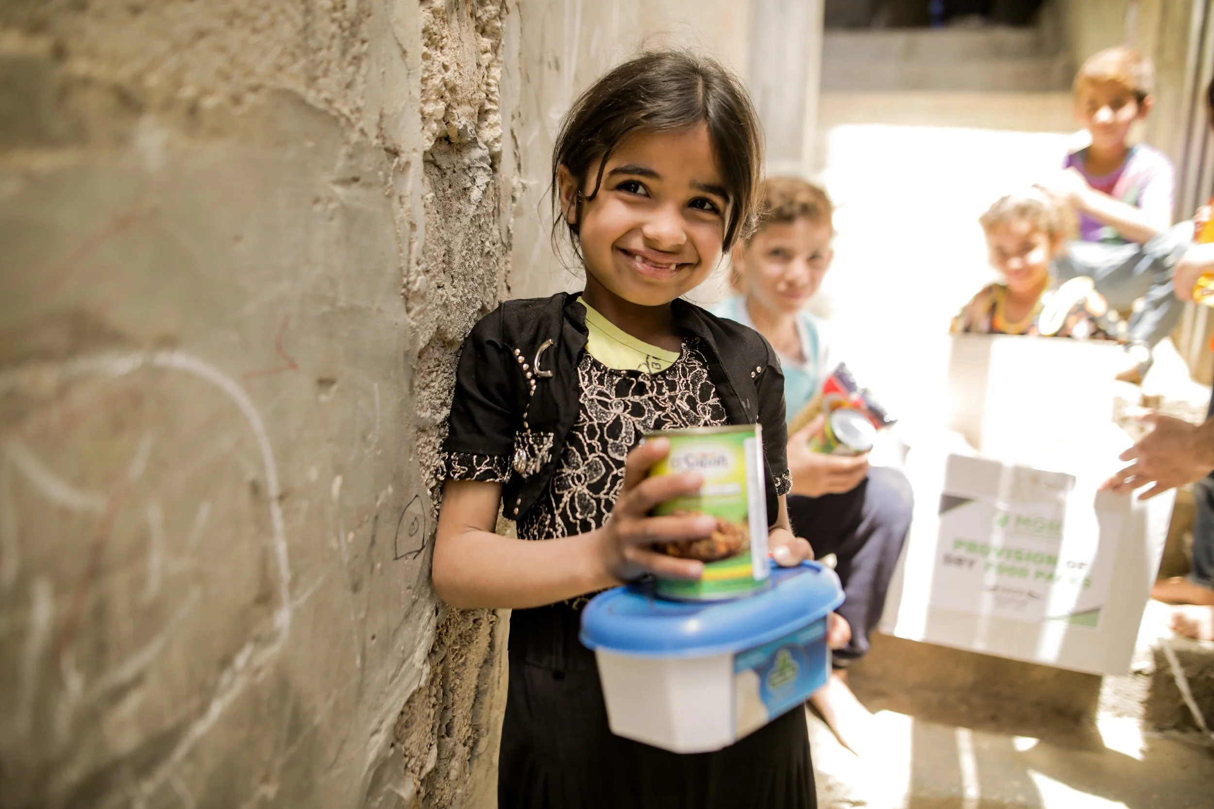 A young girl smiling while holding a canned food and a plastic container, surrounded by other children and adults in an outdoor setting with a stone wall and sunlight.