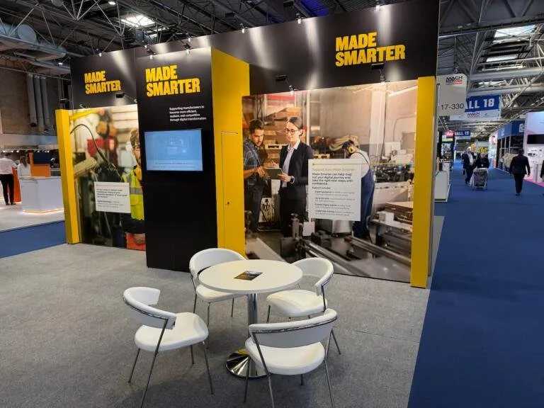 Trade show booth with yellow and black Design, labeled 'Made Smarter', featuring a large poster of people working with machinery, and a small seating area with a round table and four white chairs.