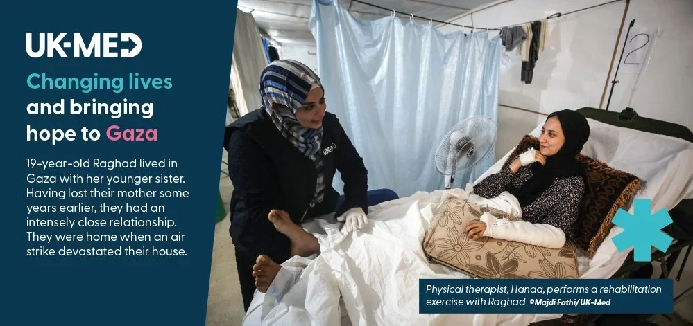 A woman in a hospital bed talking to a worker in a blue jacket and hijab. The woman is lying on her side, with a pillow behind her, while the worker stands beside her holding the patient's hand. The room has a fan, laundry hanging, and a curtain in t
