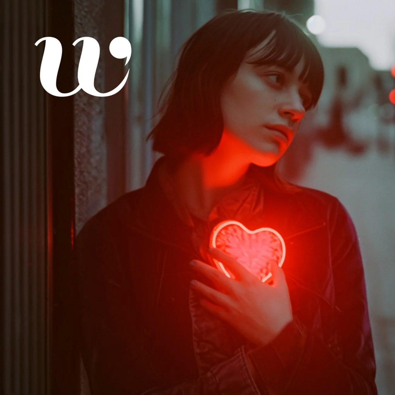 A woman with dark hair and bangs wearing a leather jacket stands against a blurred city street background at dusk. She holds her hand over her chest, which glows with a red neon heart.