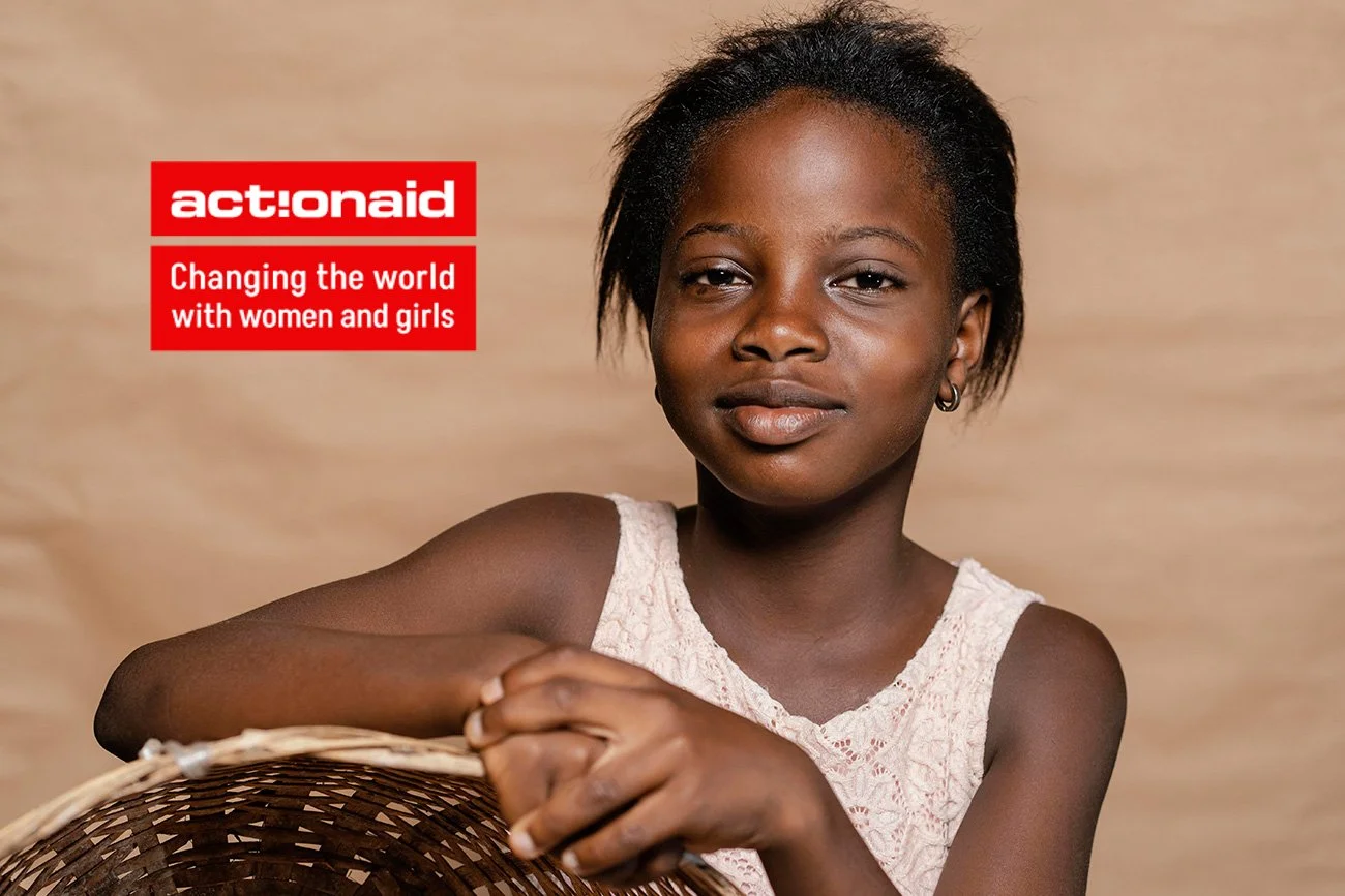 A young girl with short hair and earrings, wearing a white sleeveless top, sitting against a neutral background with a basket in front of her, and the logo of ActionAid with the text "Changing the world with women and girls".
