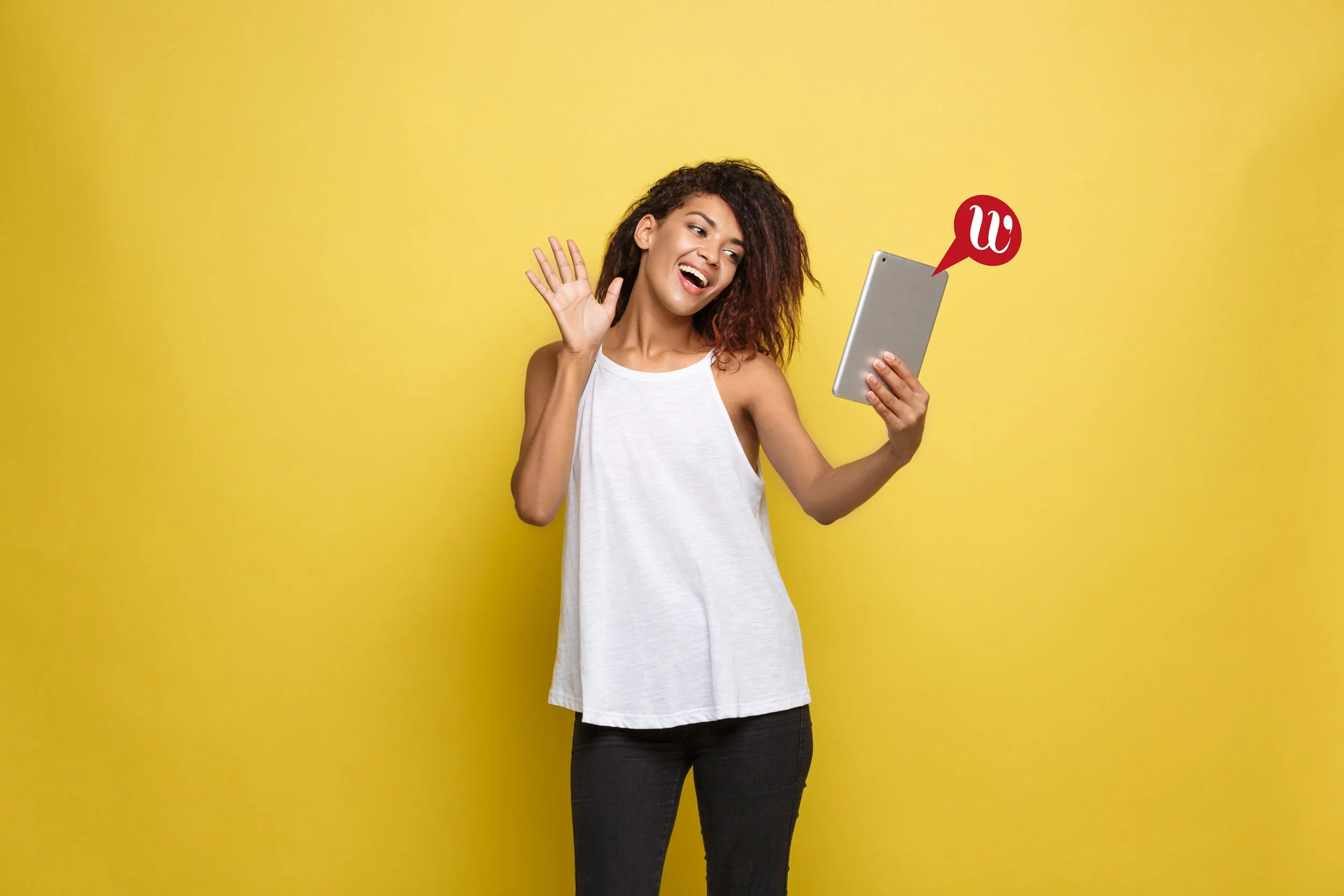 A woman with curly hair wearing a white sleeveless top and black pants is smiling and waving while holding a tablet. A red speech bubble with a white 'W' is near the tablet.