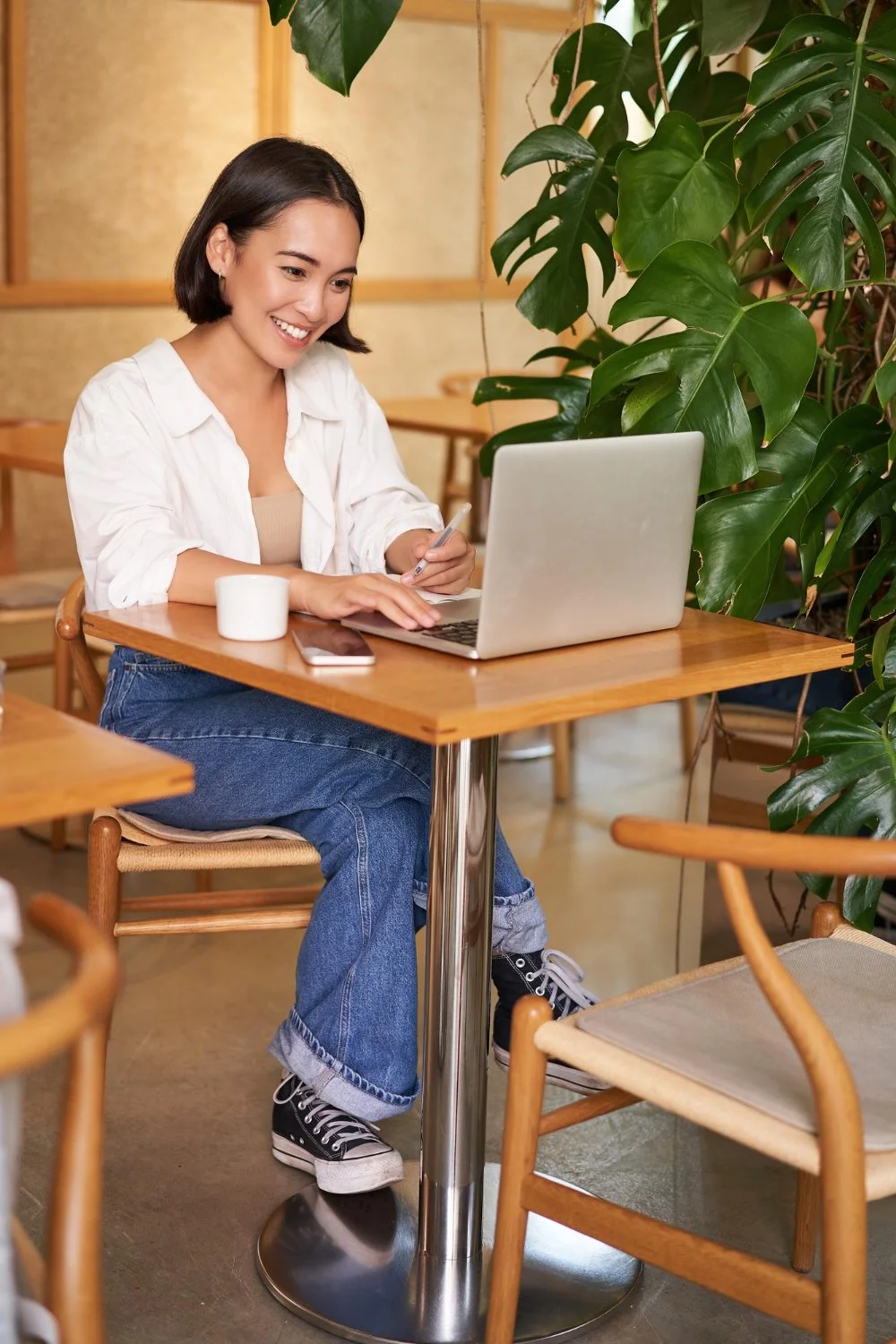 A woman with short dark hair, wearing a white shirt and jeans, sitting at a wooden table in a cafe, smiling at her laptop with a large green plant next to her.
