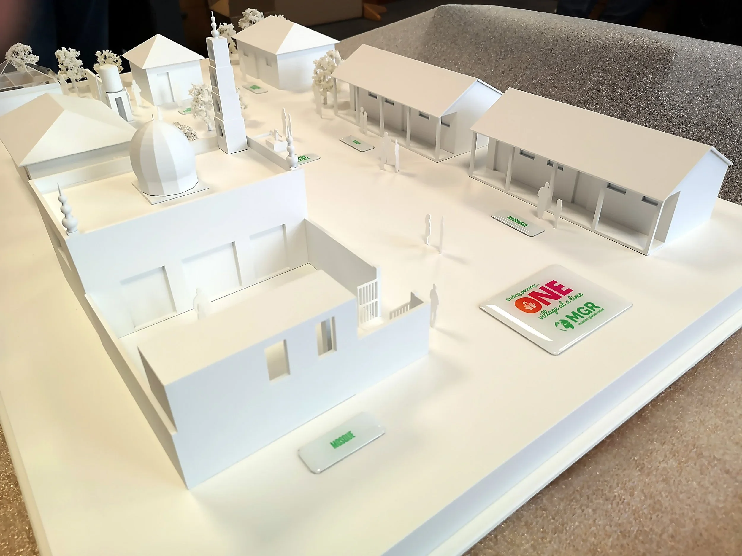 A white architectural model of a community with various buildings, trees, and miniature human figures, including a domed structure and a clock tower, on a table with labels indicating different areas or projects.