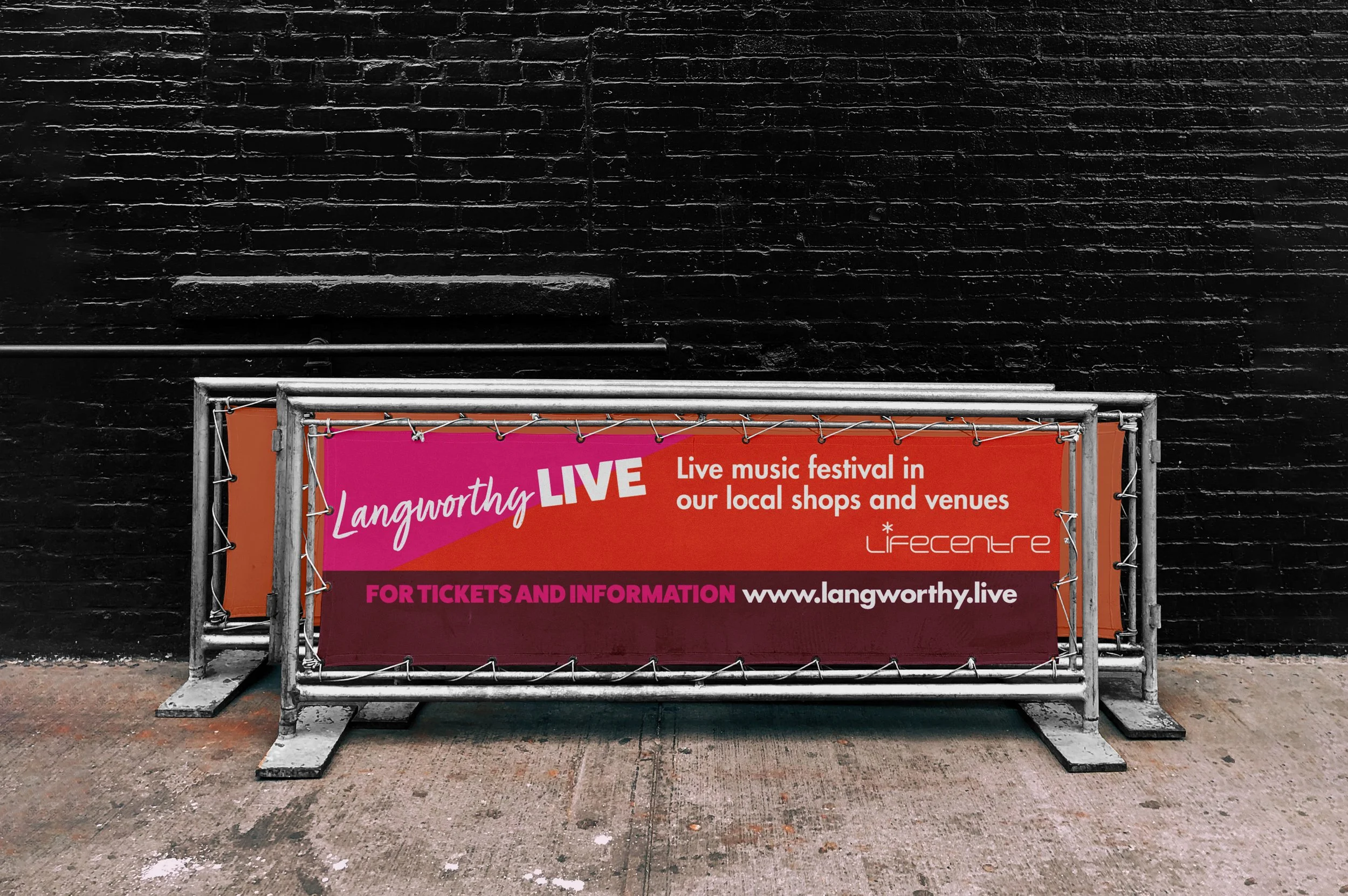 A street banner advertising a live music festival called Langworthy LIVE, happening in local shops and venues, with ticket and information website www.langworthy.live, supported by Lifecentre, displayed on a metal barricade against a black brick wall.