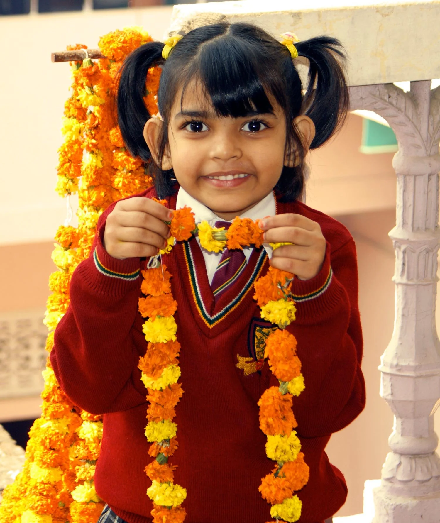 A young girl with pigtails, wearing a school uniform and red sweater, smiling and holding a marigold flower garland, standing near a floral decoration.