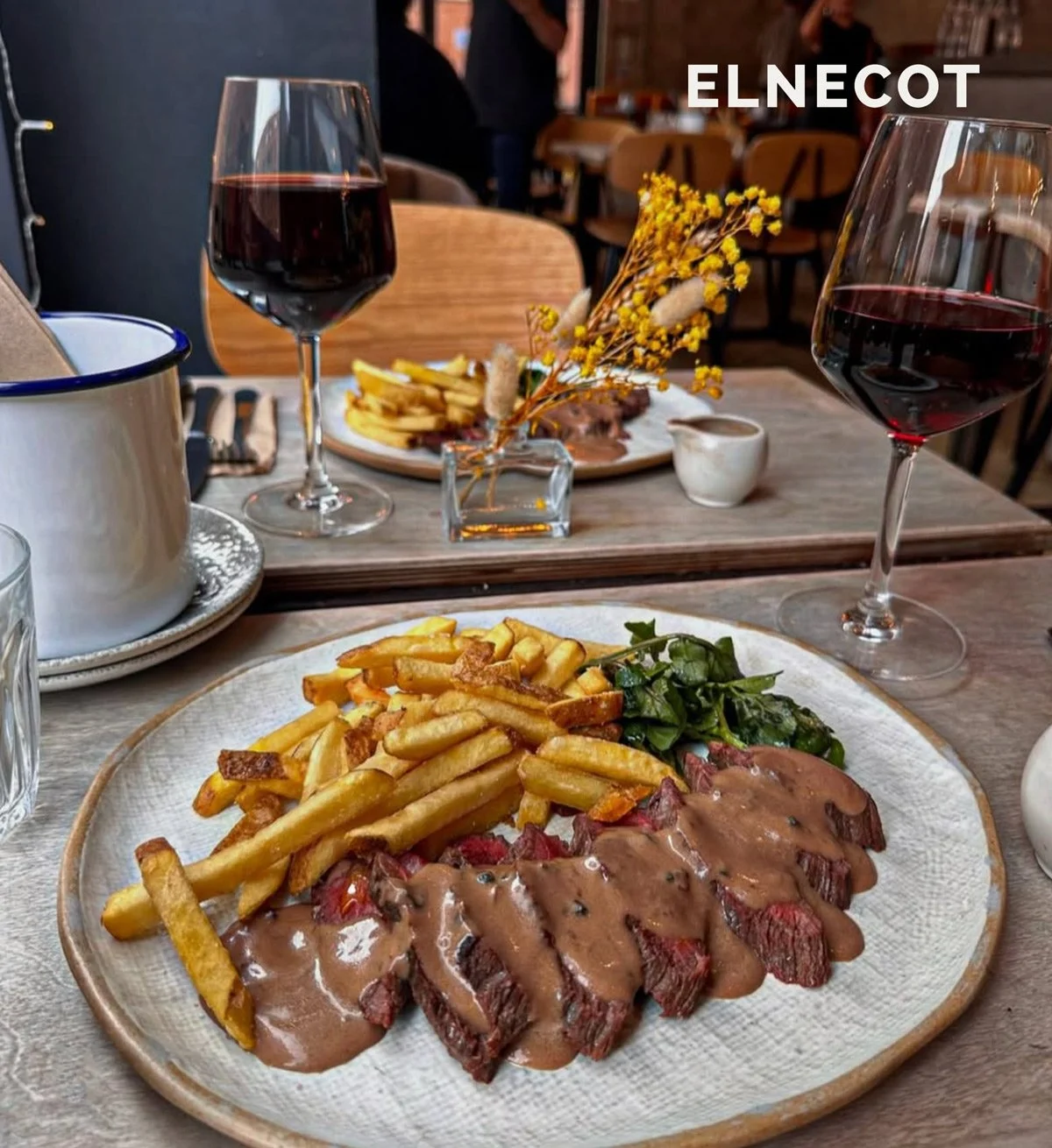 A table set with two glasses of red wine, a plate of French fries and sautéed greens, and a plate of sliced steak with gravy. In the background, there is a small vase with yellow flowers, a white cup, and a second plate of food.