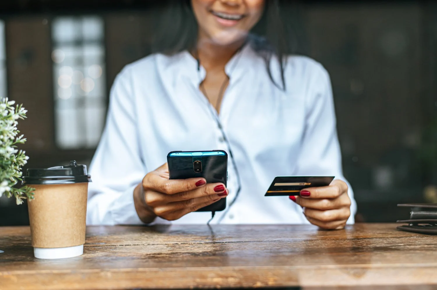 Woman sitting at a wooden table, holding a smartphone and a credit card, with a takeout coffee cup and a small plant nearby.