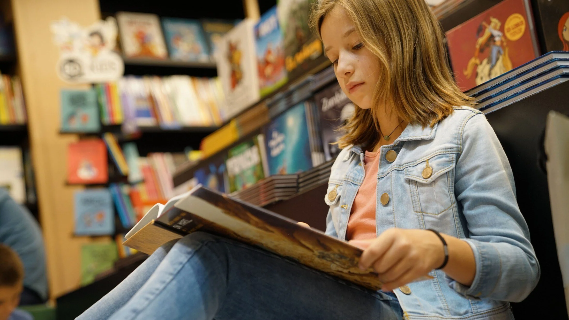 A young girl in a denim jacket reading a book in a bookstore.