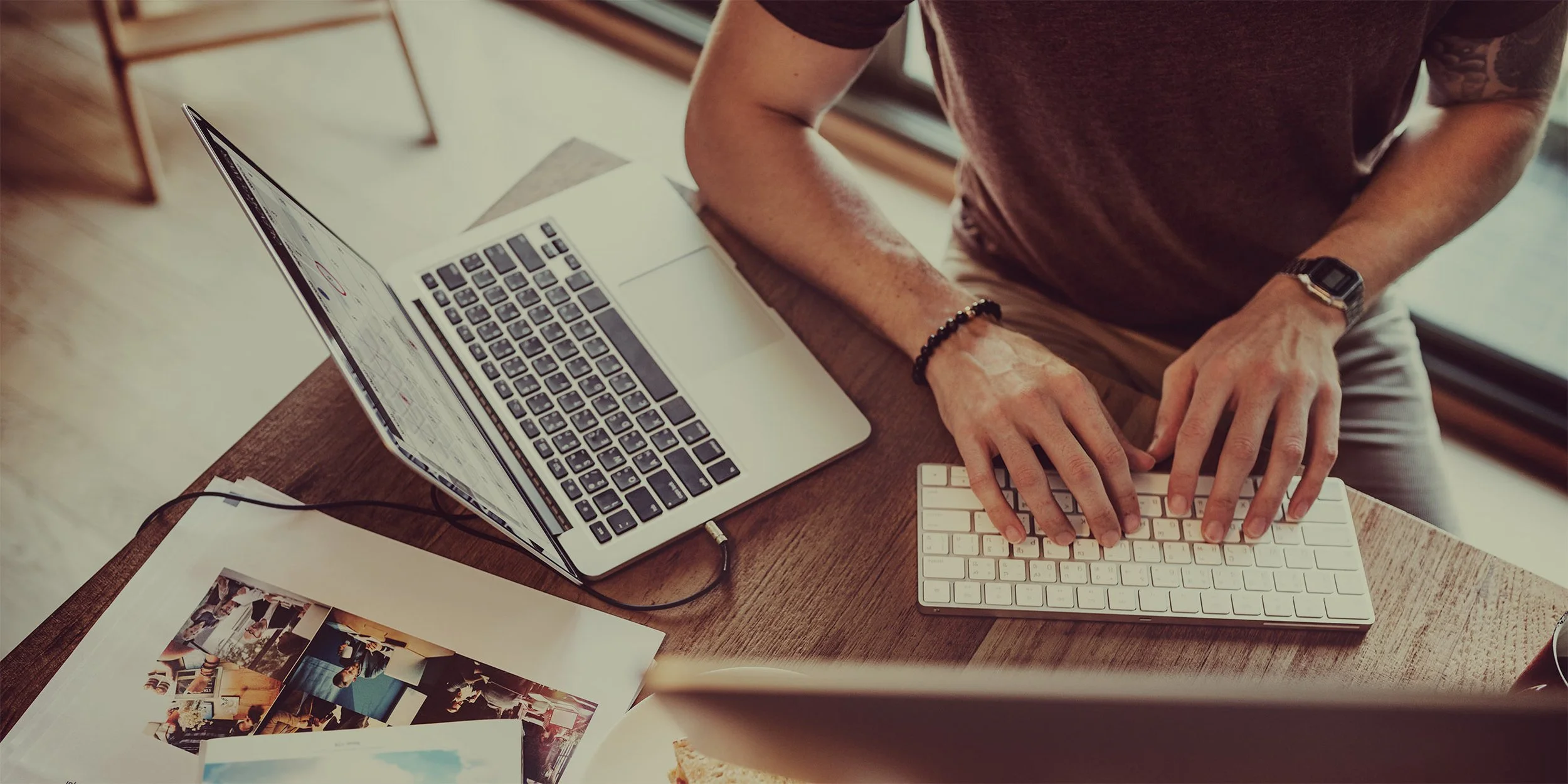Person working on a laptop and keyboard at a wooden desk, with printed photos and a coffee mug nearby.