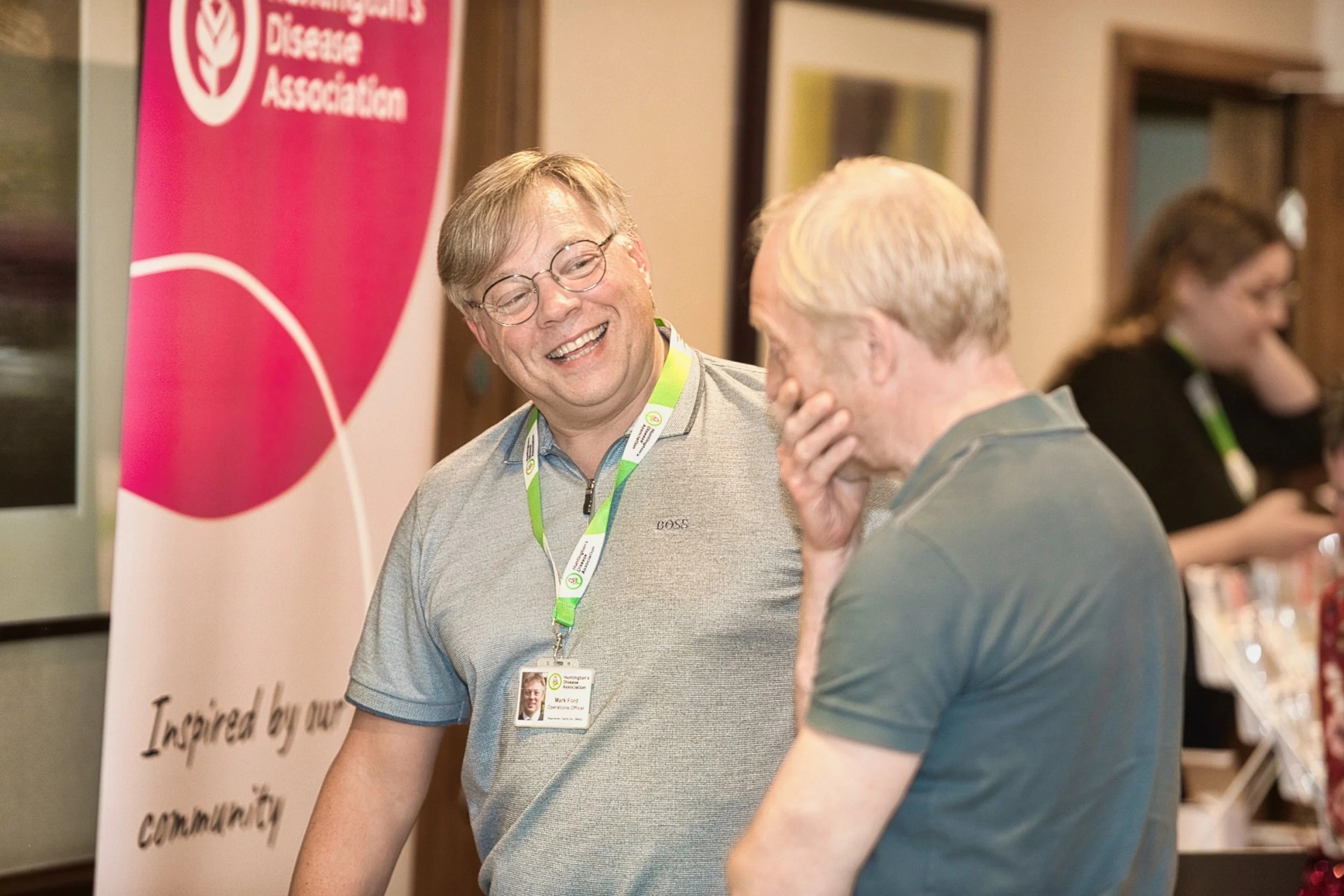 Two men at a breast cancer awareness event, one wearing glasses and a gray polo shirt, smiling and talking to the other who has his hand on his face. A pink and white banner for the Huntington's Disease Association is in the background.