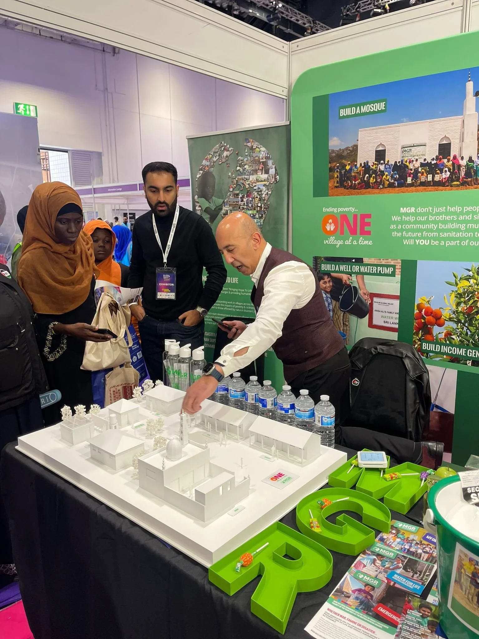 A man is explaining a scale model of a community center at an exhibit booth, with a group of diverse people observing. The booth has a large green sign promoting community projects like building a mosque, well, and water pump, with informational posters and bottles of water on the table.