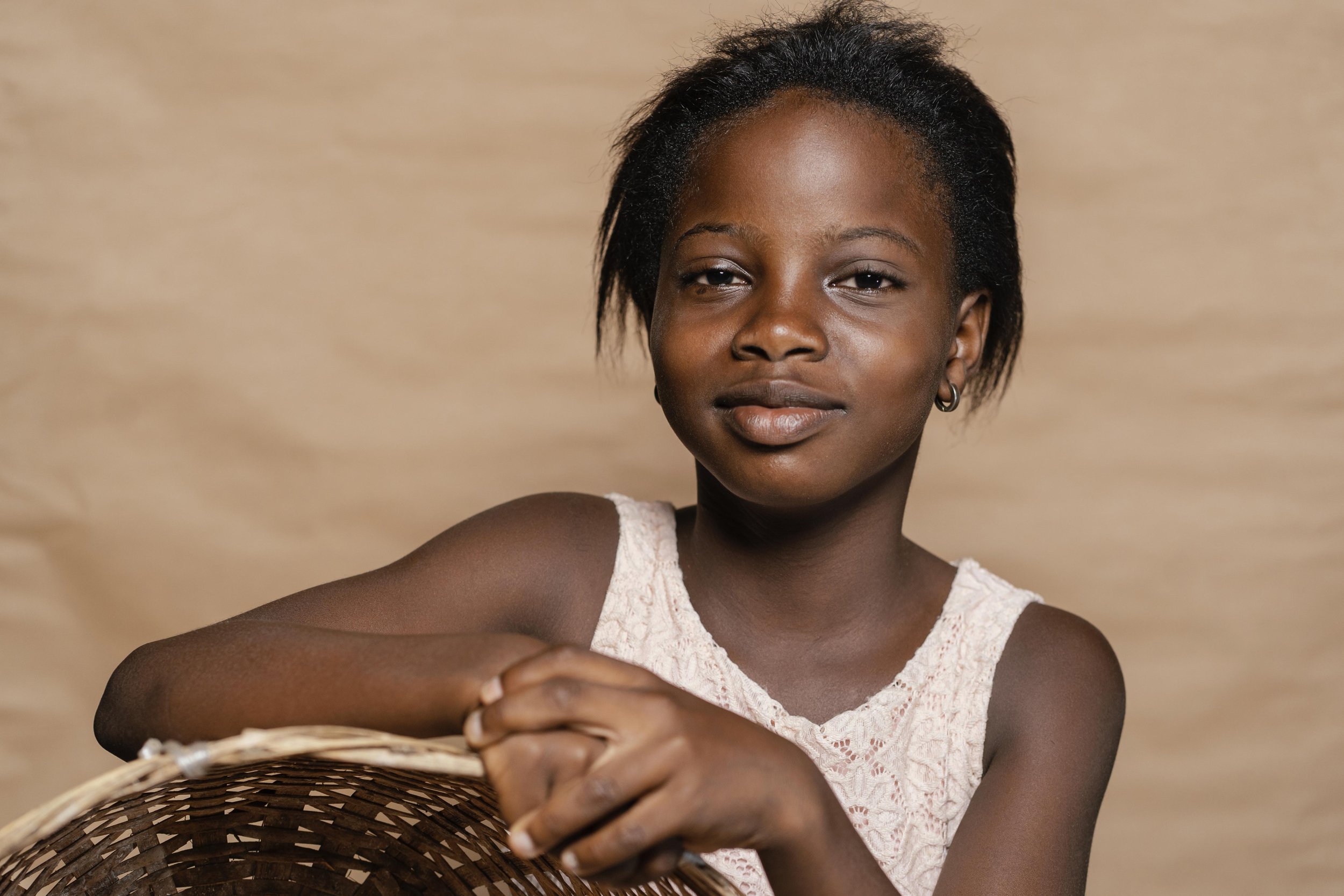 Portrait of a young woman with short black hair wearing a white lace sleeveless top, sitting with her arms crossed on a wicker surface against a beige background.