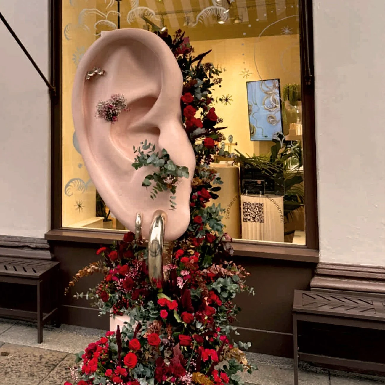 Decorative display featuring a large pink ear-shaped sculpture adorned with earrings, flowers, and greenery, outside a shop window with Christmas decorations inside.