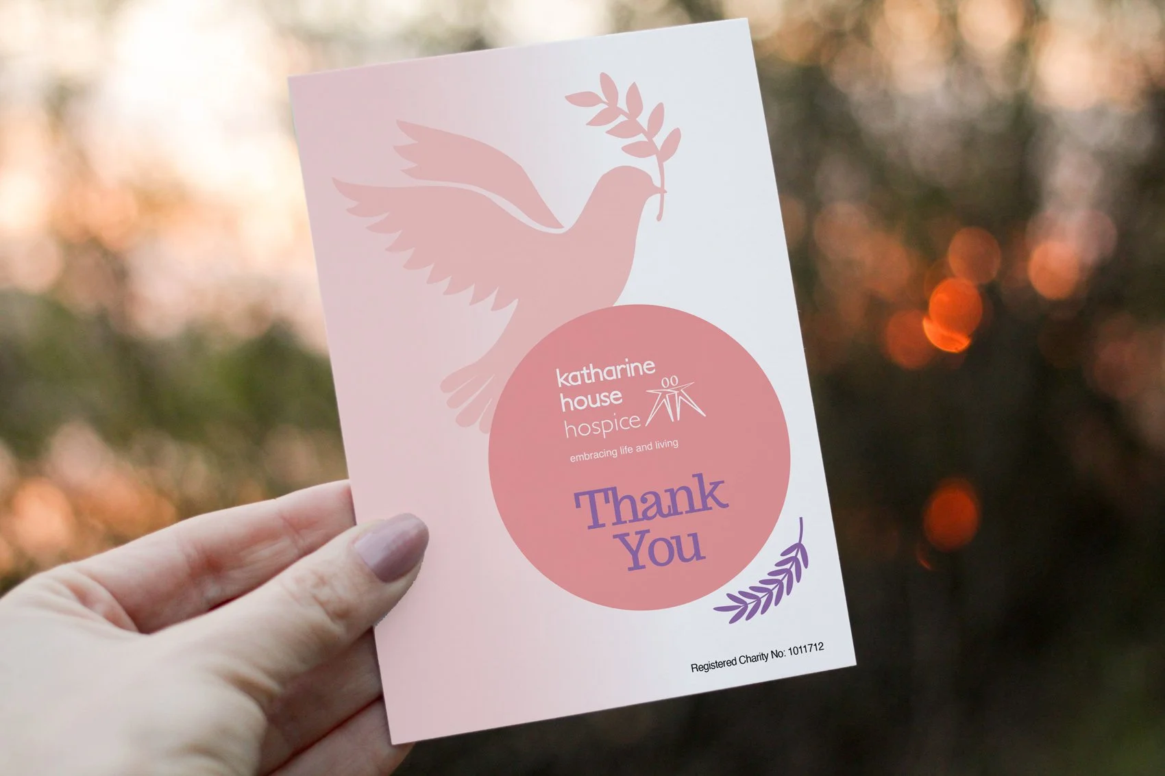 Hand holding a pink thank you card from Katharine House Hospice with a dove holding an olive branch and decorative leaves, set against a blurred outdoor background with warm bokeh lights.
