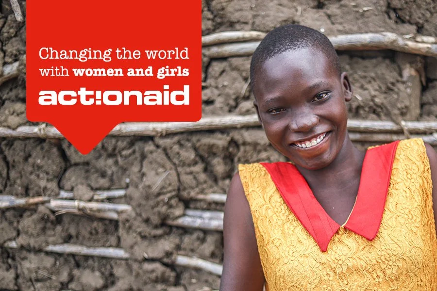 A young girl with short hair smiling and wearing a yellow dress with red accents, standing in front of a mud wall with straw reinforcement.