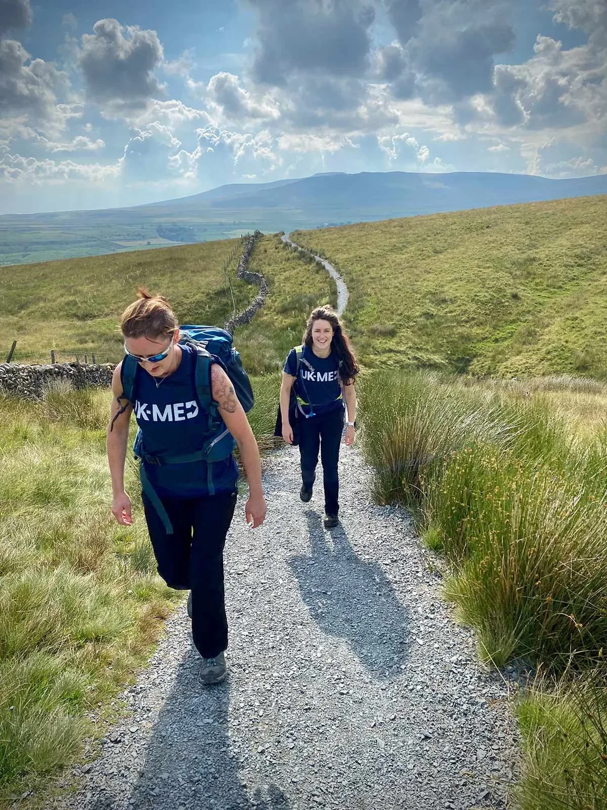 Two women hiking on a gravel trail in a green, open countryside with hills and mountains in the background under a partly cloudy sky.