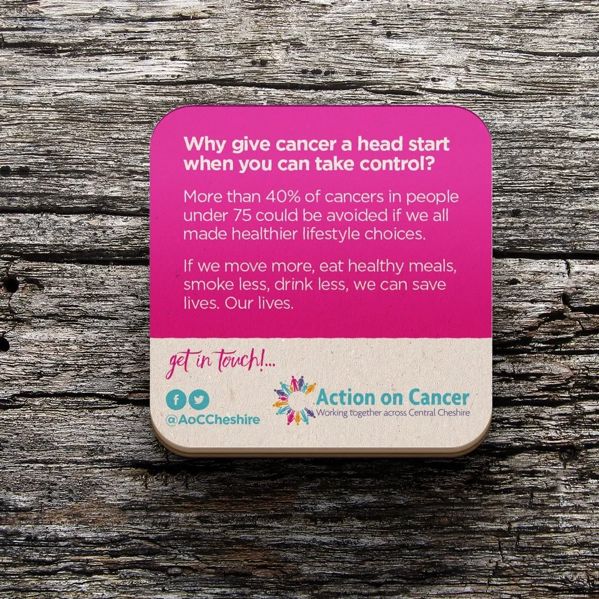 Pink and white informational card promoting healthy lifestyle choices to reduce cancer risk, with social media handles and a colorful logo at the bottom, placed on a wooden surface.