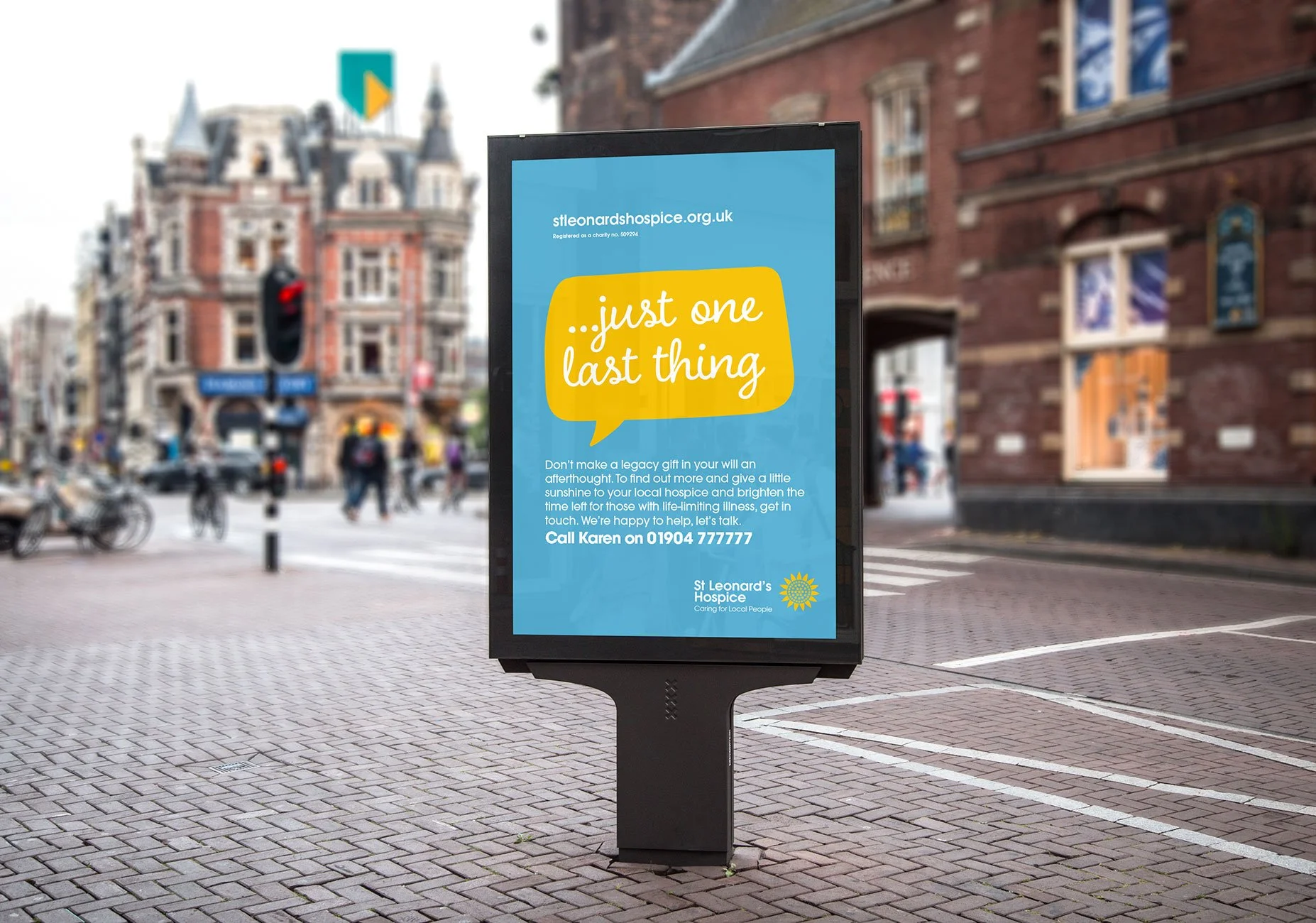 A digital billboard on a city street sidewalk promoting St Leonard's Hospice. The billboard has a blue background with a yellow speech bubble that says, '...just one last thing.' Text below encourages donations and provides a contact number, with the